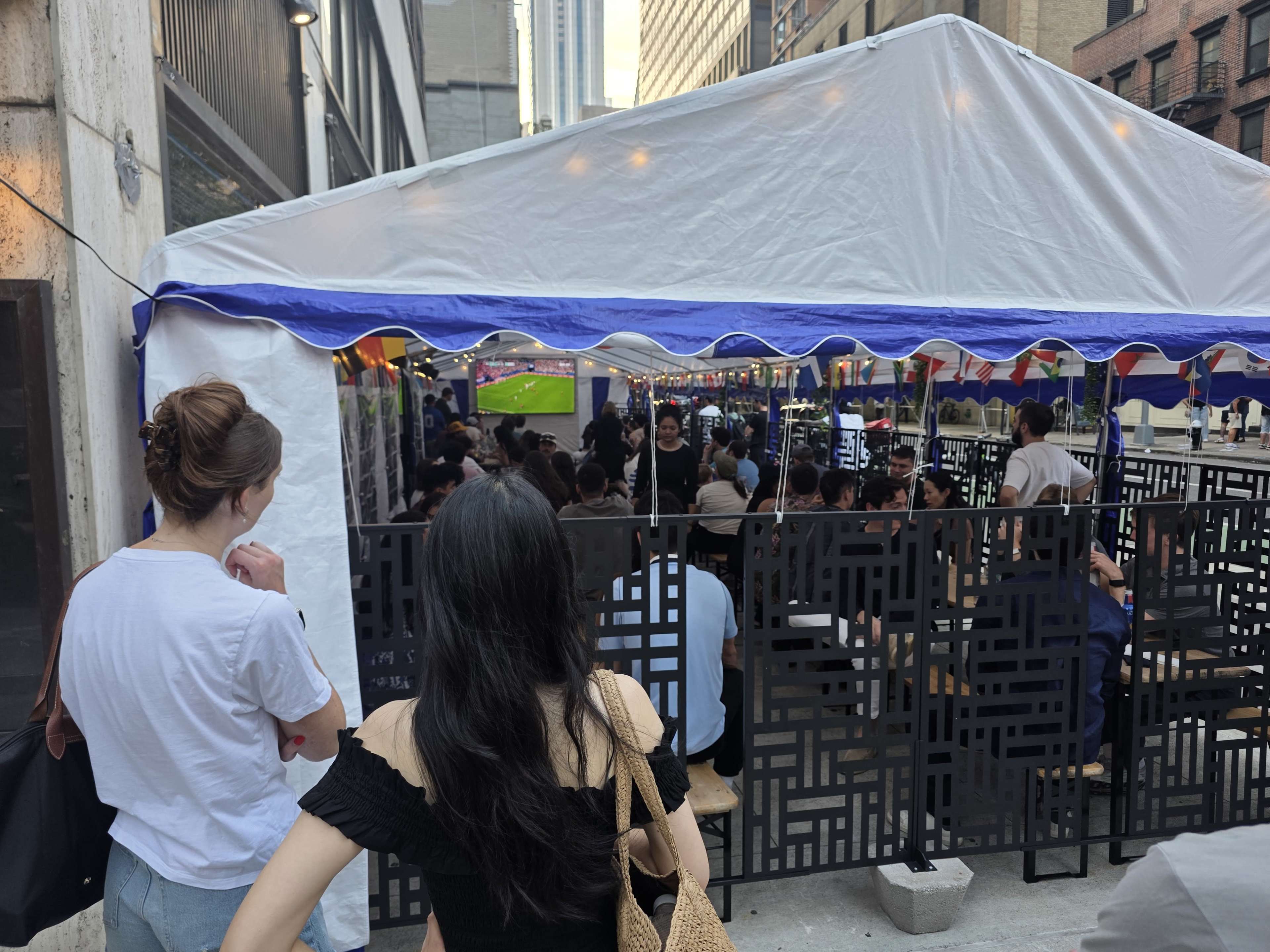 A large outdoor tent is filled with people watching a soccer game on a screen, while several spectators stand in line outside.