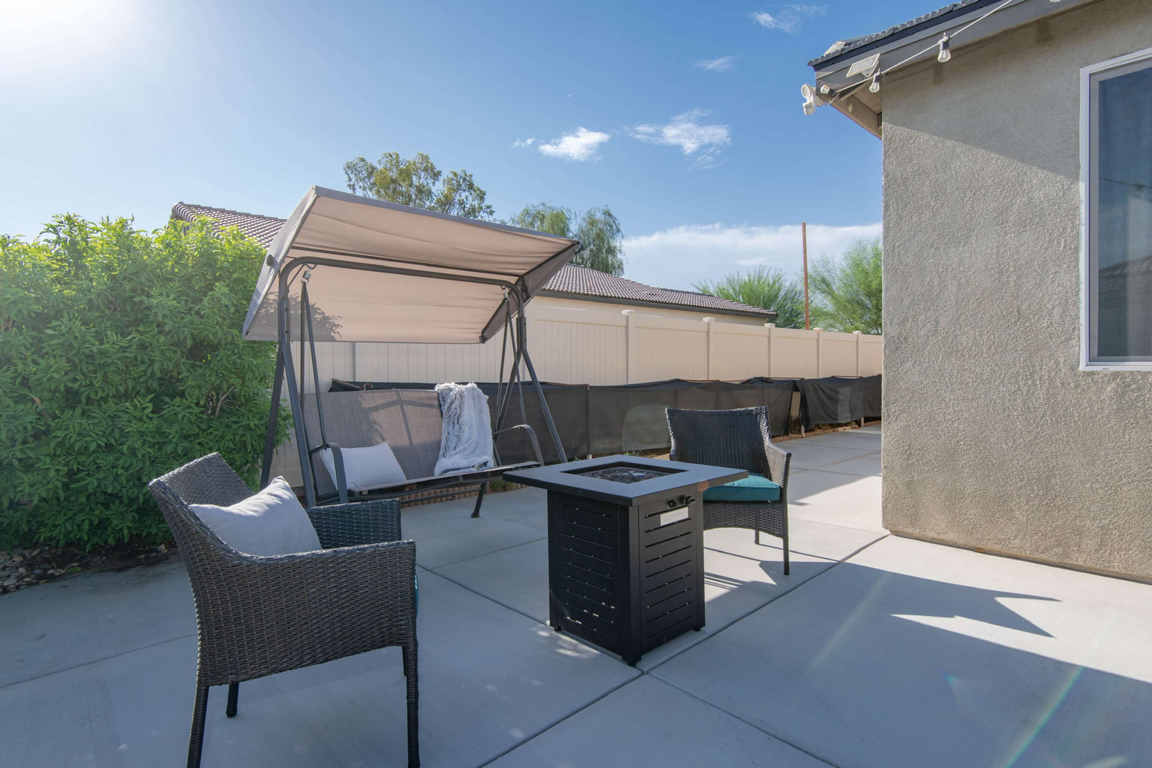 A covered swing and two chairs surround a rectangular fire pit on a concrete patio in a backyard.