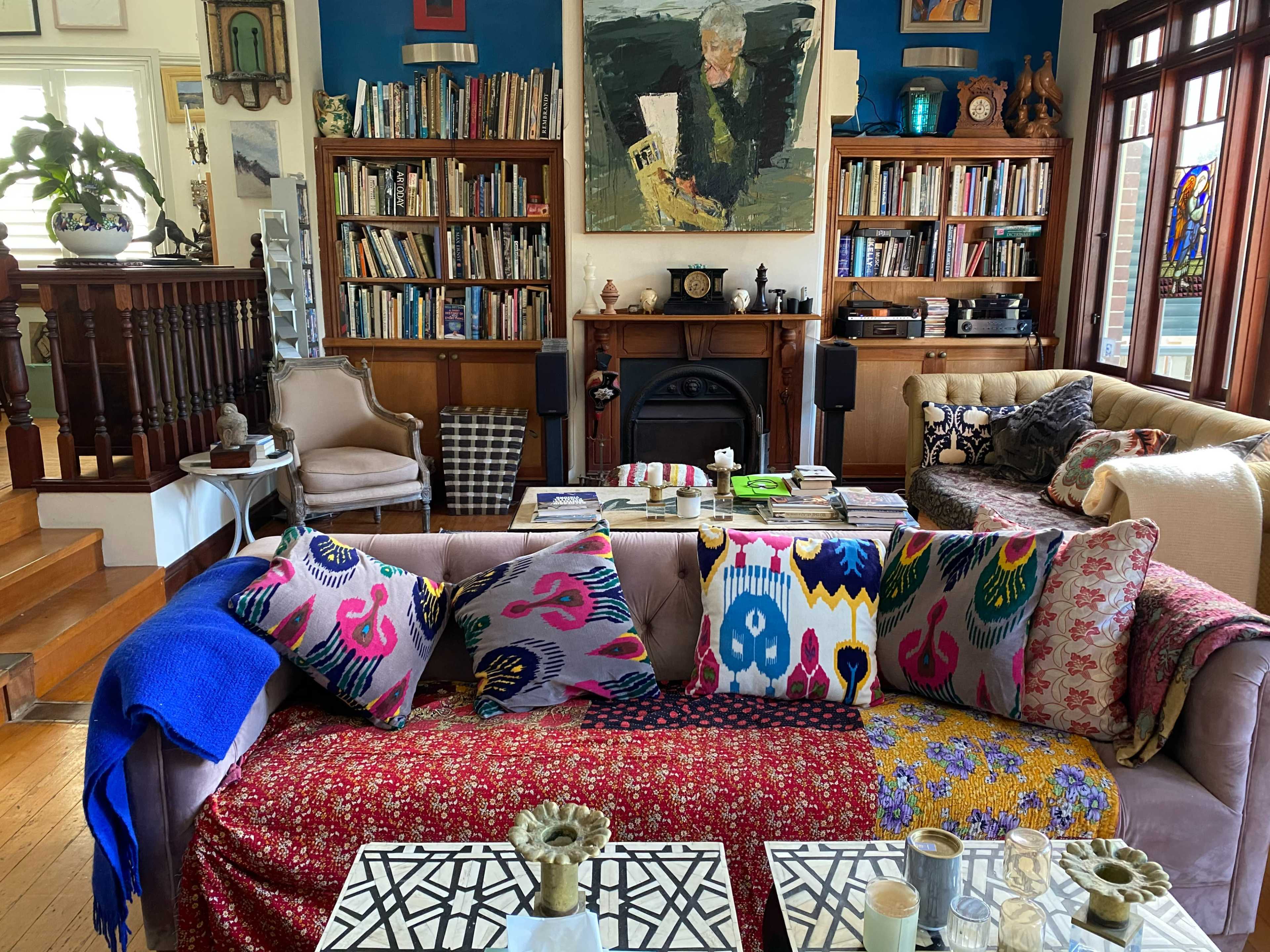 The image shows a cozy living room with a mix of bookshelves, colorful cushions on sofas, a decorative fireplace, and a variety of artworks on the walls.