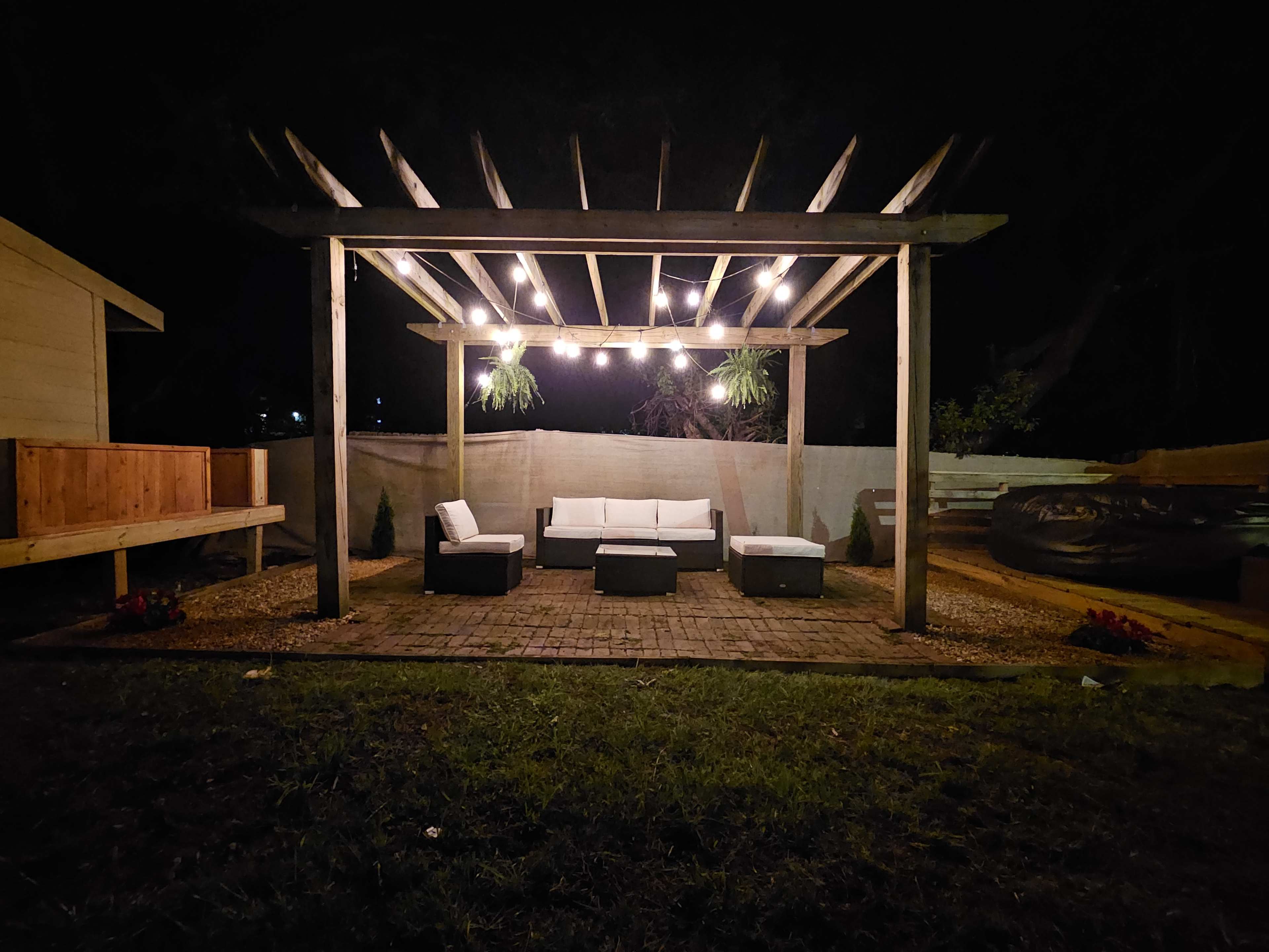 A patio area at night features a wooden pergola with hanging lights, surrounded by furniture including a white sectional sofa and black chairs, set on a stone-paved floor.