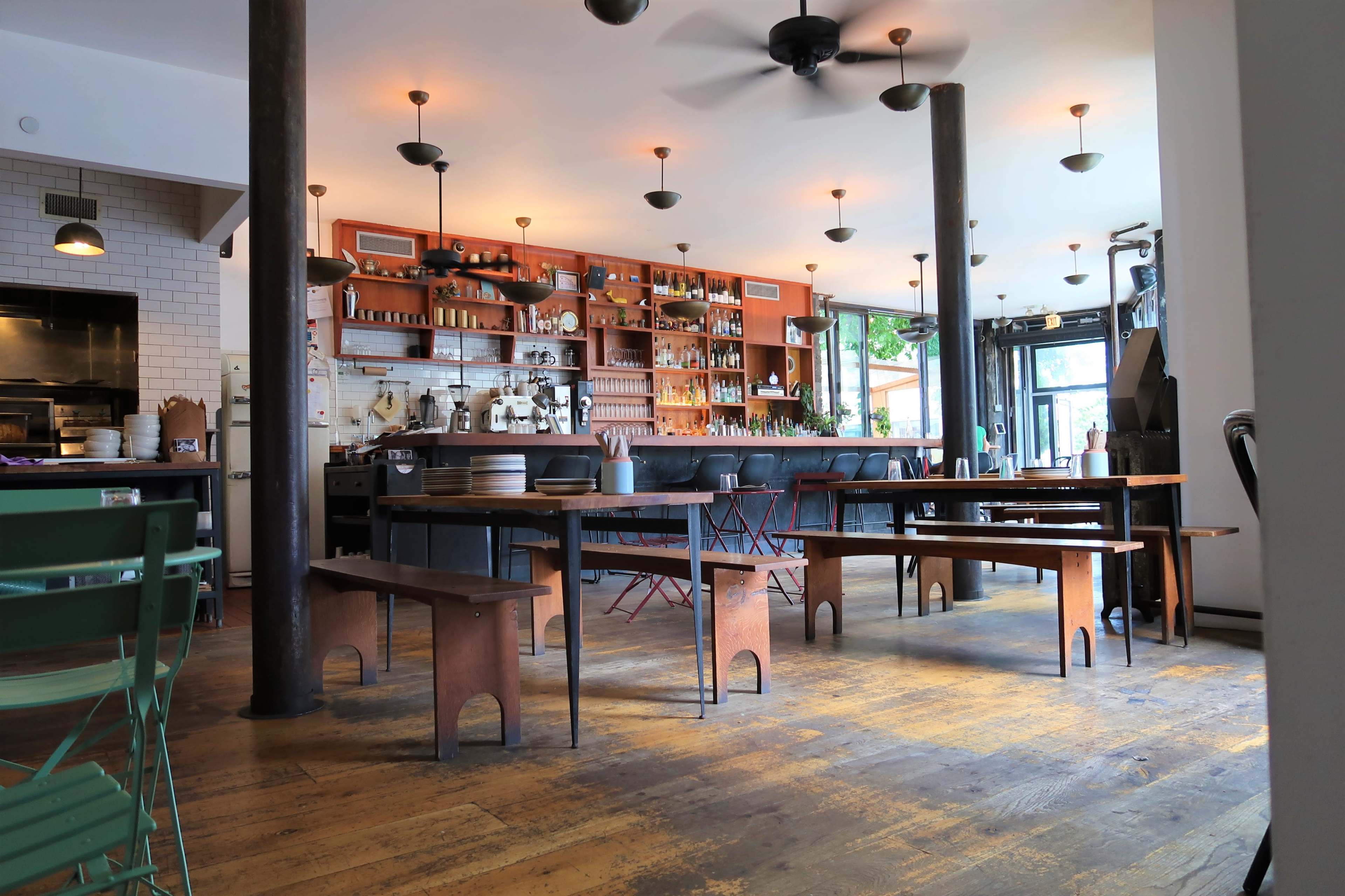 A spacious café interior with wooden flooring, a bar area with a variety of bottles, and several tables arranged throughout the room.