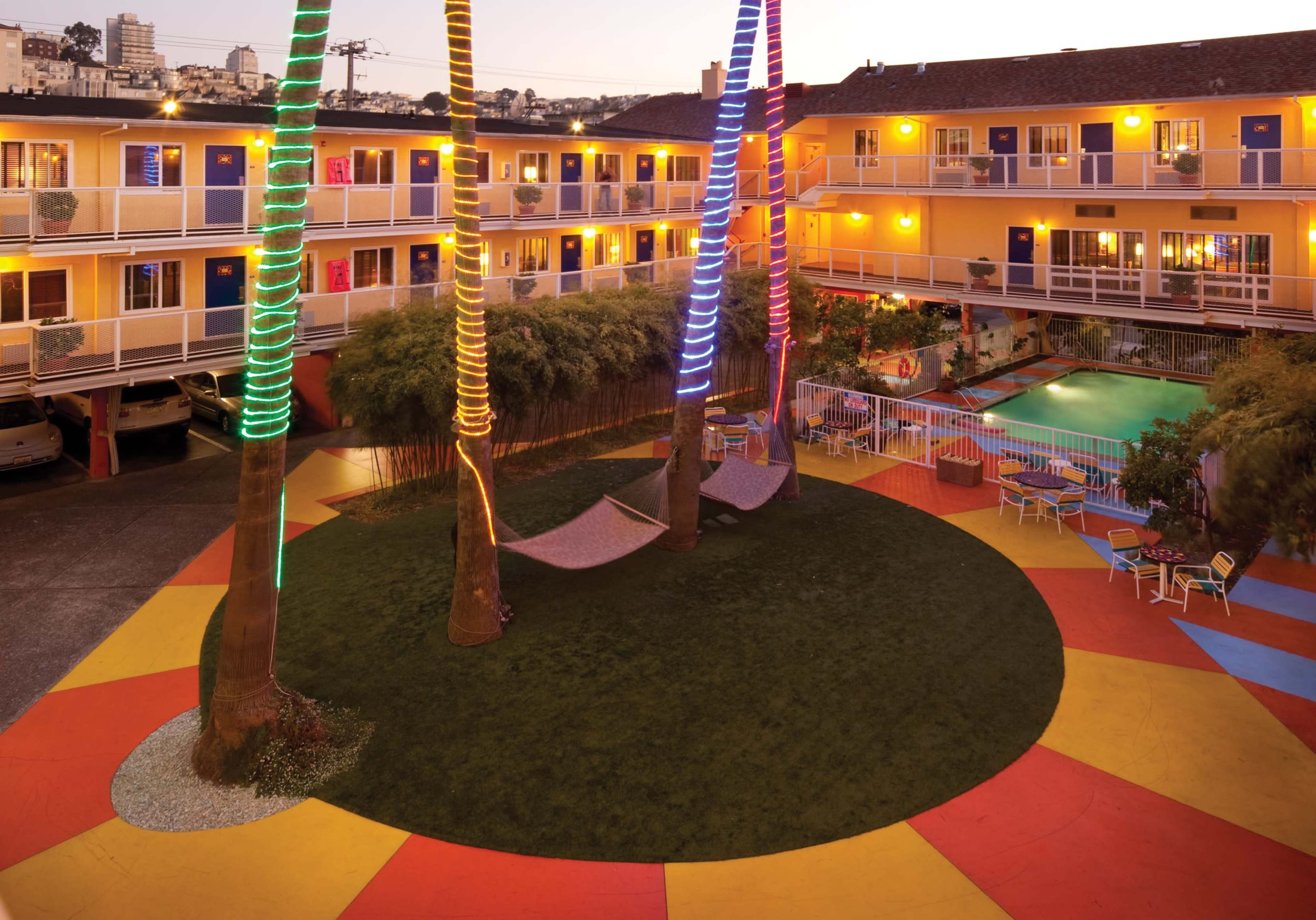 A colorful motel courtyard features palm trees wrapped in lights, two hammocks, and a swimming pool surrounded by vibrant tiles.