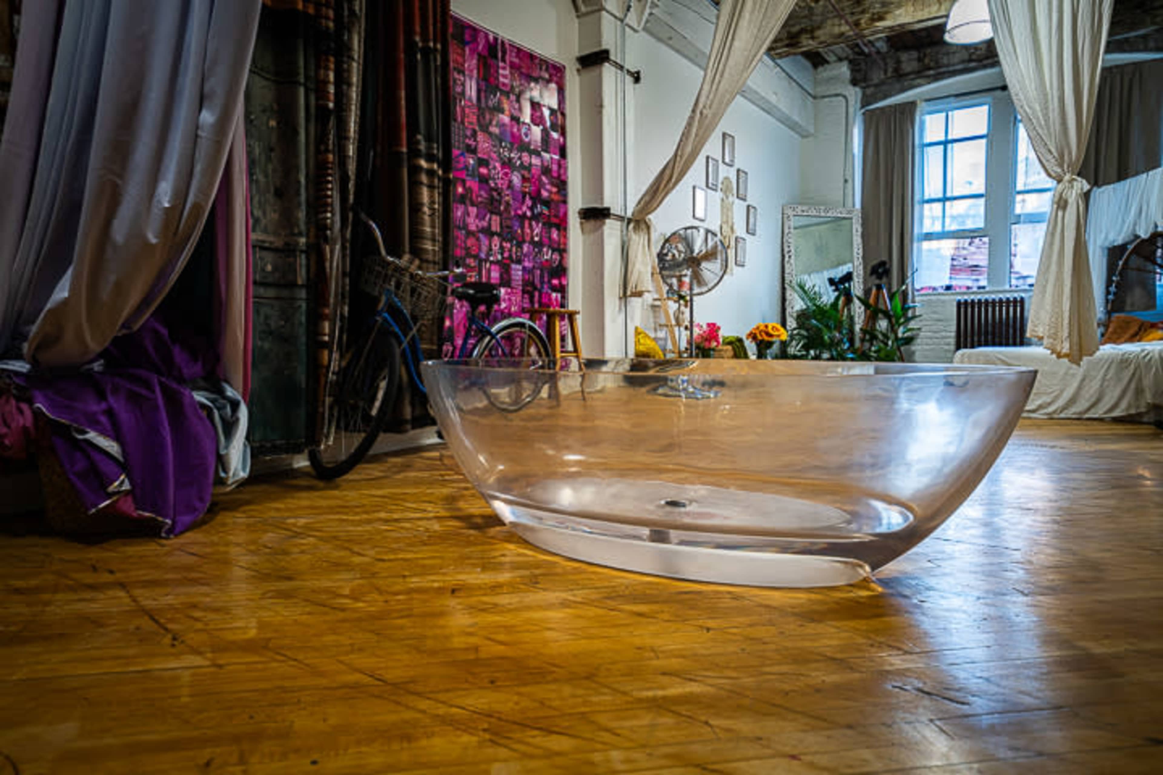 A large, transparent acrylic bathtub is placed on a wooden floor inside a stylishly decorated room with plants and a bicycle in the background.