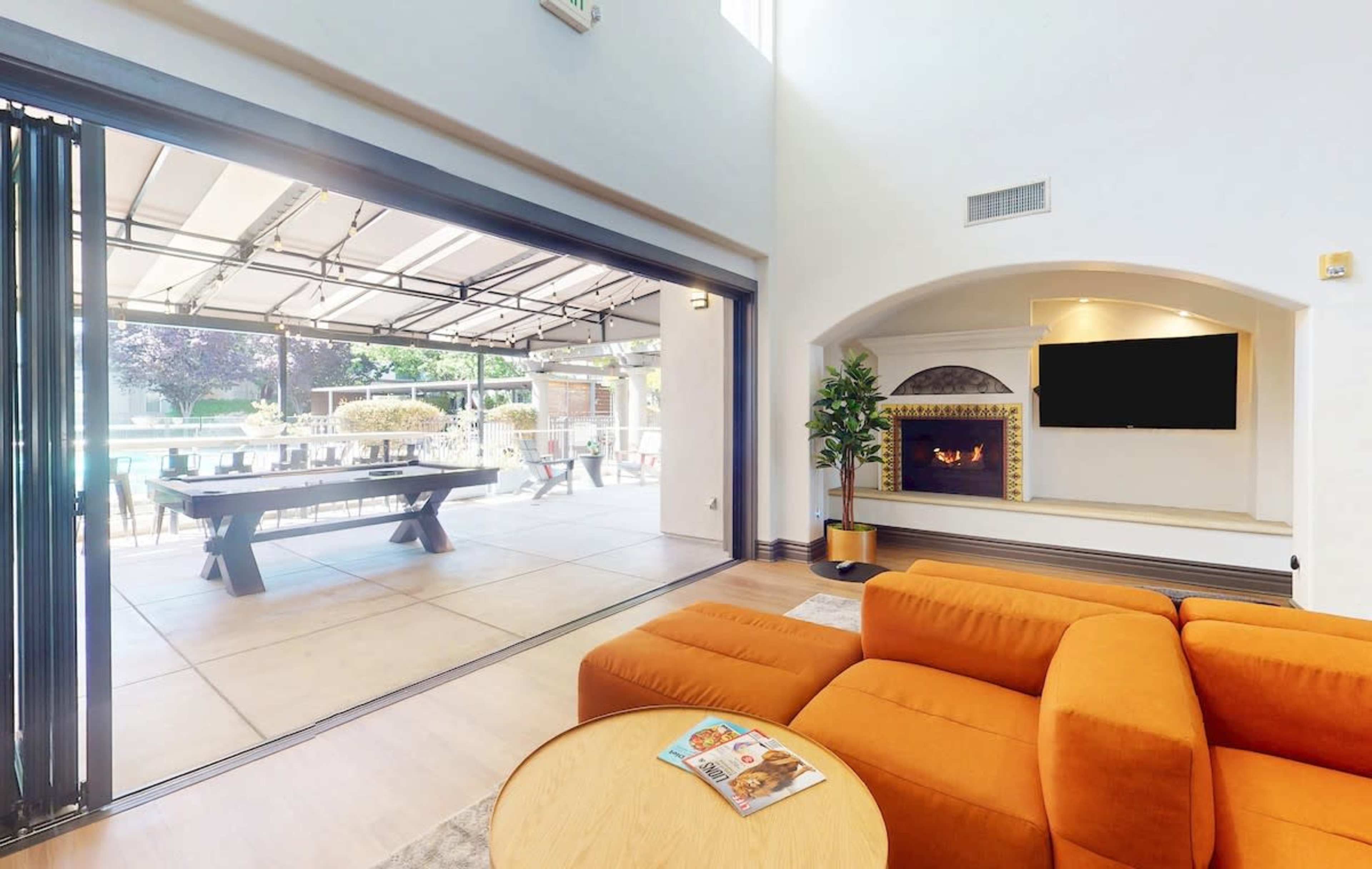 The image shows a spacious living area featuring an orange sectional sofa, a coffee table, and a large open door leading to an outdoor patio with a pool and dining area.