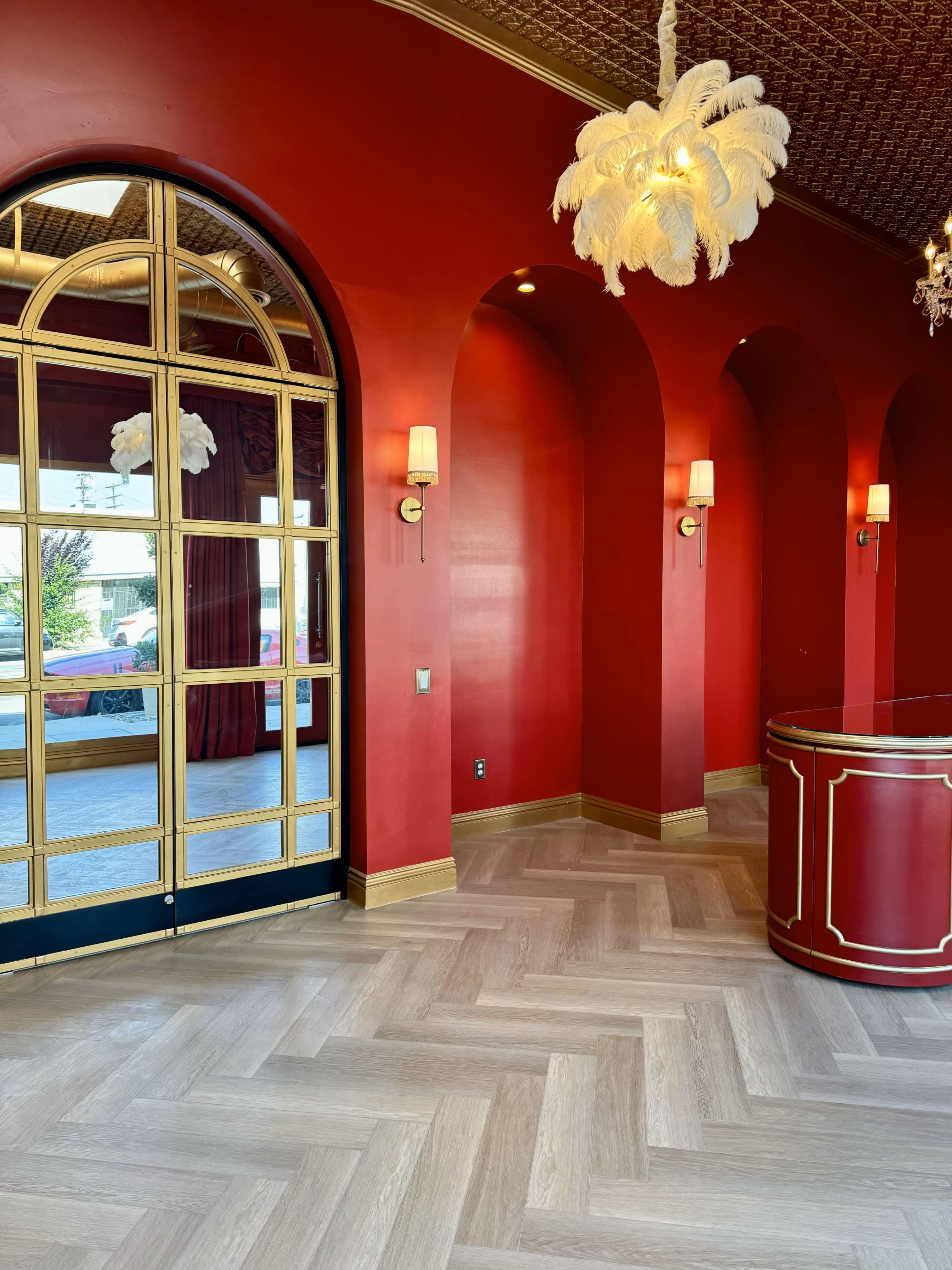 A red-walled interior with arched doorways, a mirrored wall, and a decorative reception desk in a herringbone-patterned wooden floor.