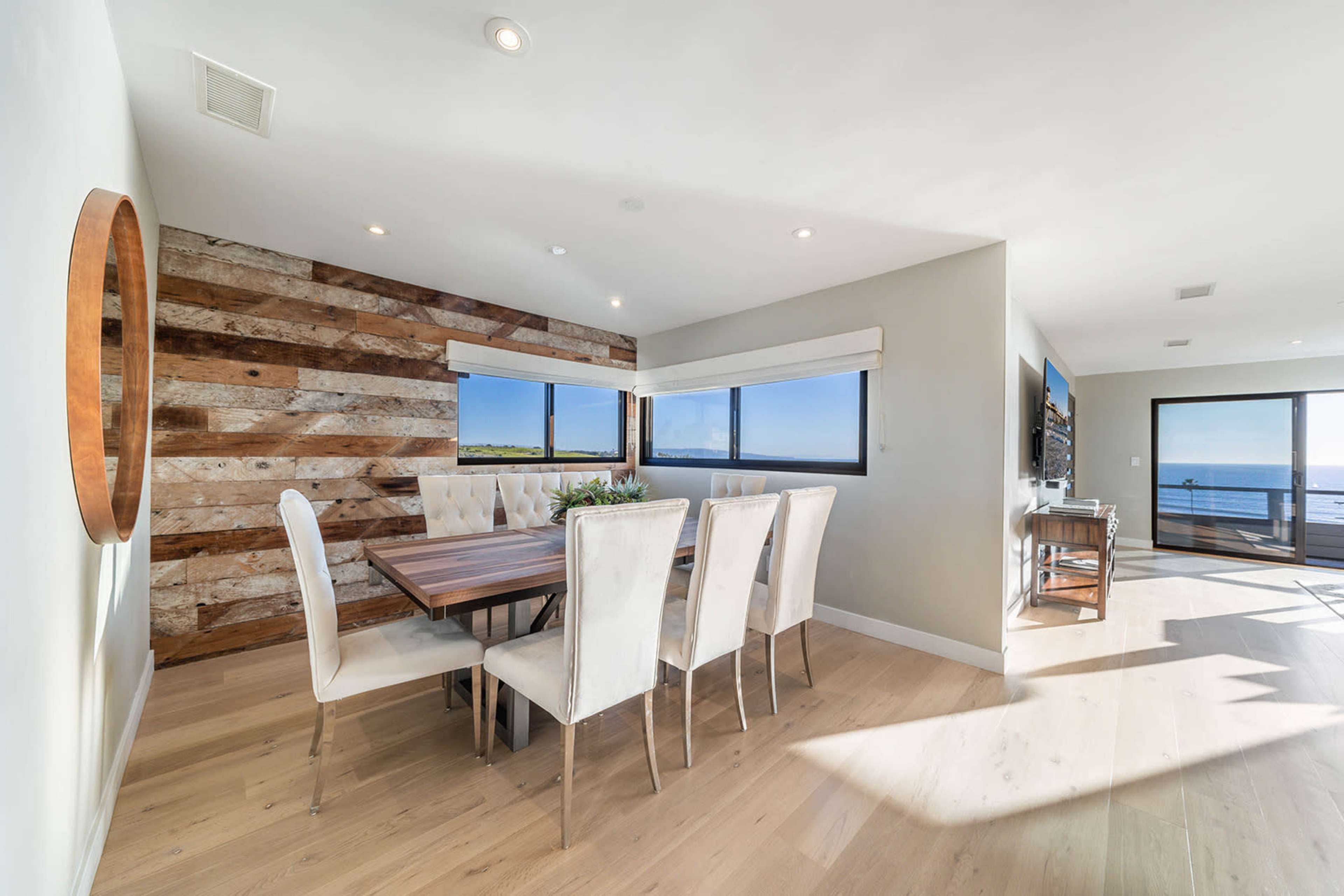 A modern dining area features a wooden table surrounded by white upholstered chairs, with large windows offering a view of the ocean.