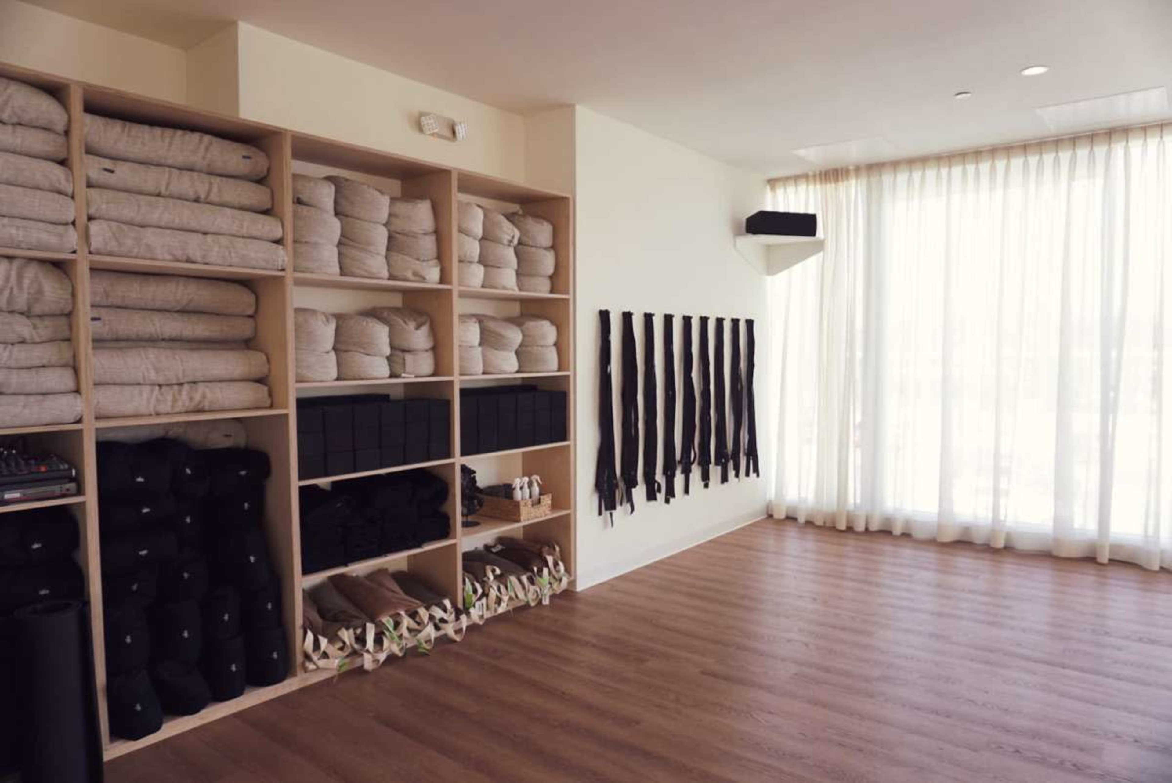 The image shows a minimalist yoga studio with neatly arranged shelves containing folded blankets, blocks, and belts, alongside a spacious area with wooden flooring and large windows.