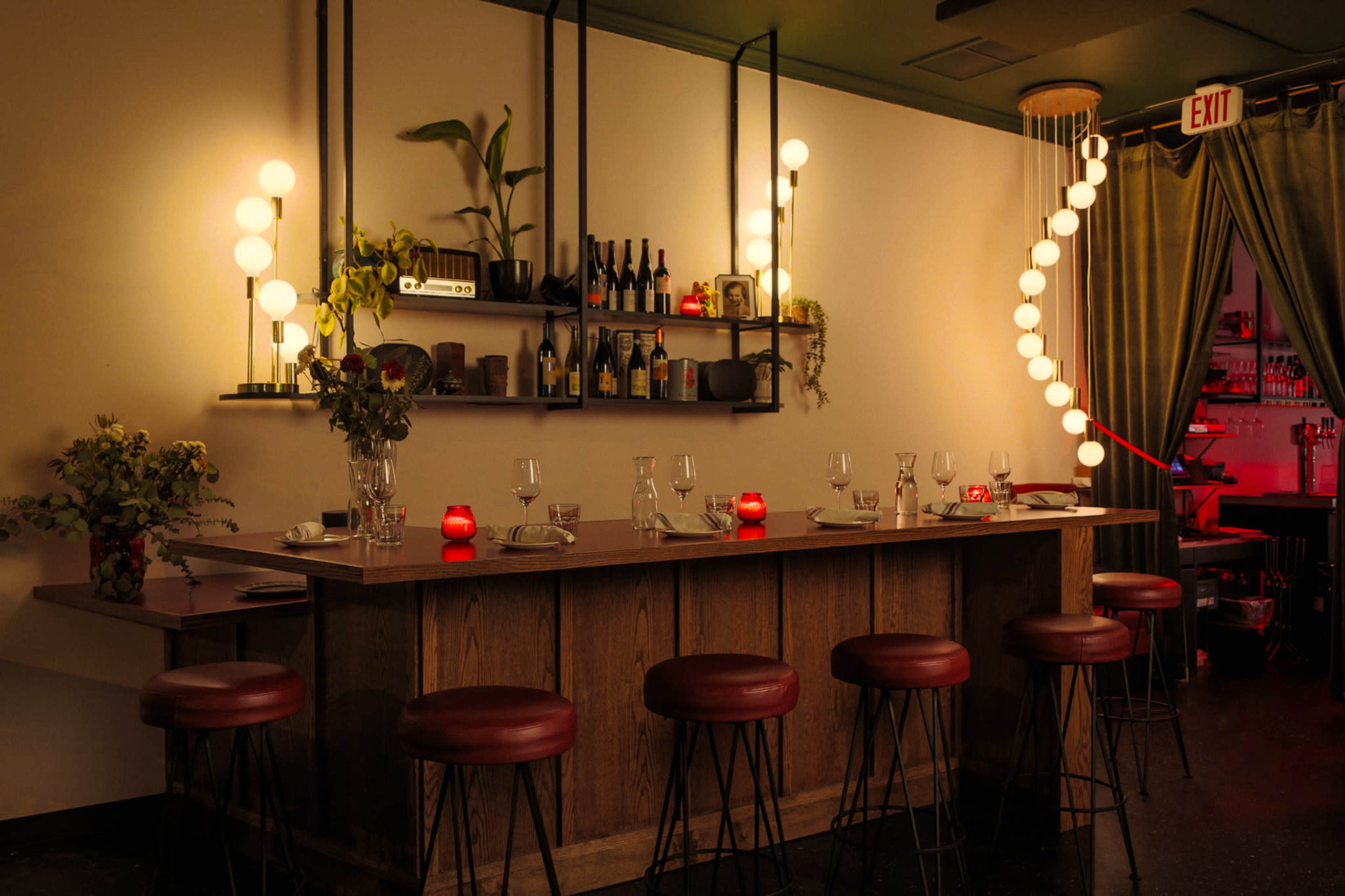 The image shows a dimly lit bar area with a wooden counter, red stools, and decorative lighting, featuring shelves with bottles and plants.