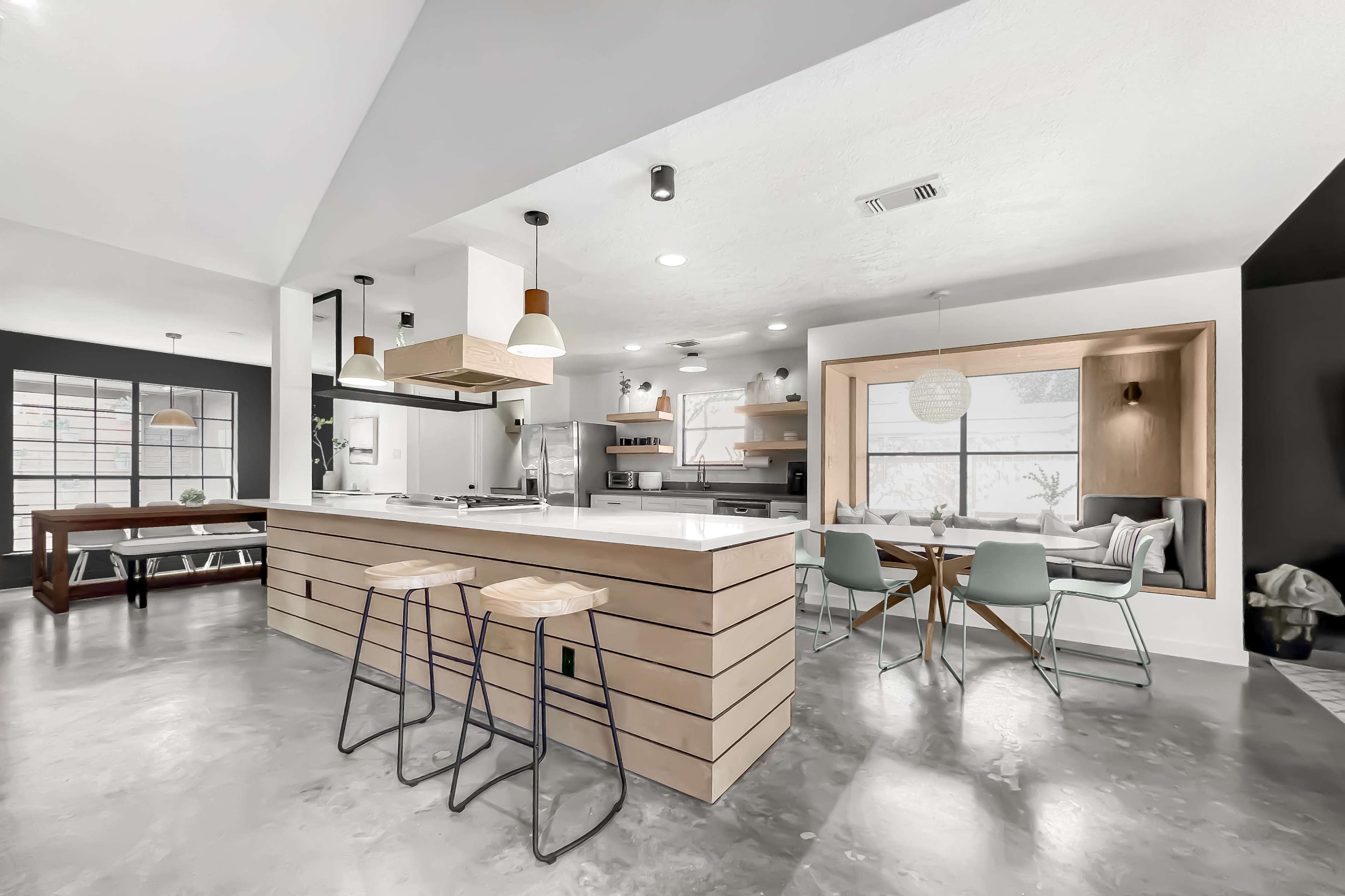 A modern kitchen featuring an island with wooden paneling, bar stools, and a dining area with a table and chairs, all illuminated by pendant lights.
