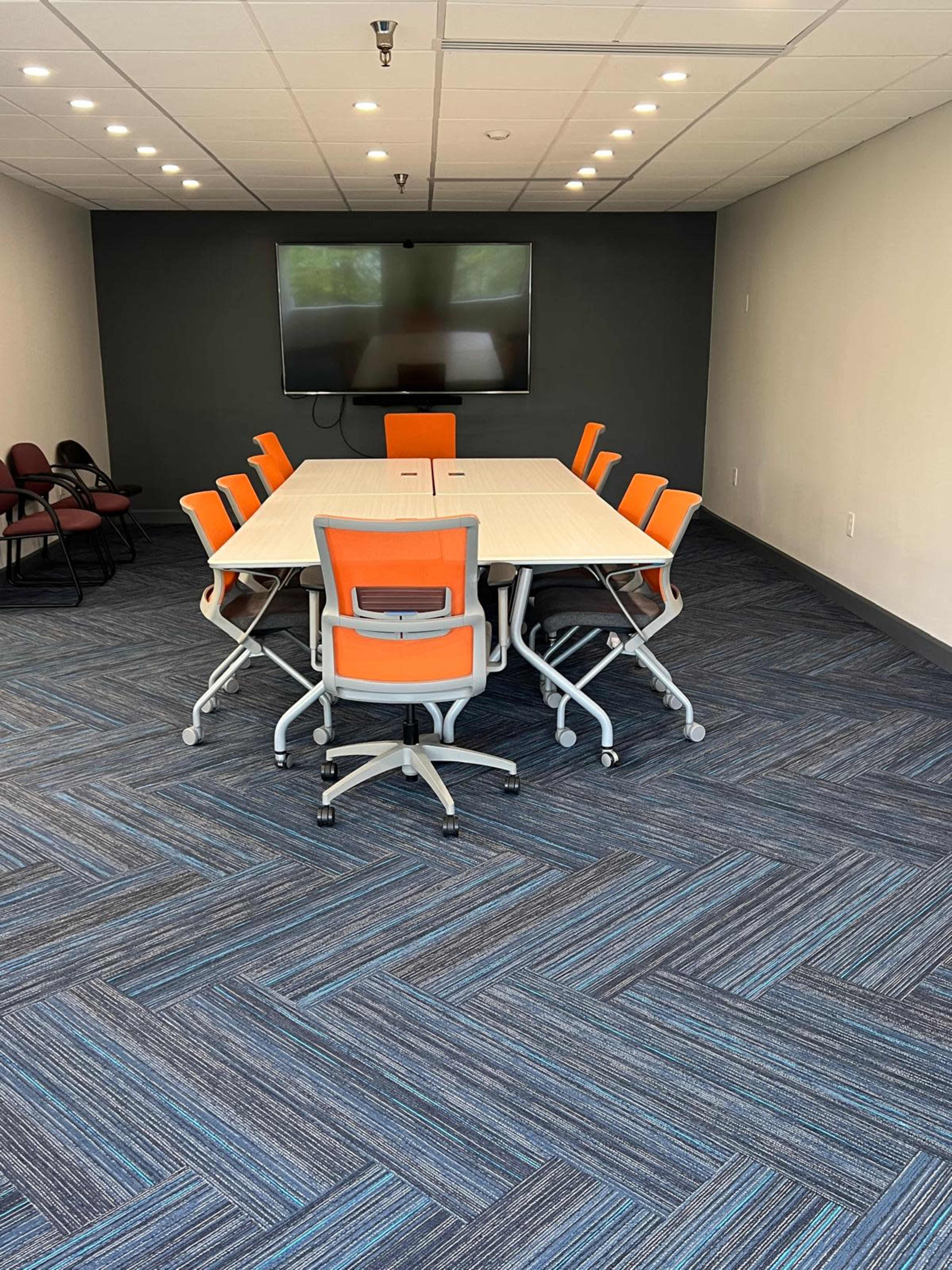 A modern conference room features a large, rectangular table surrounded by eight orange and gray office chairs, with a wall-mounted television at one end.