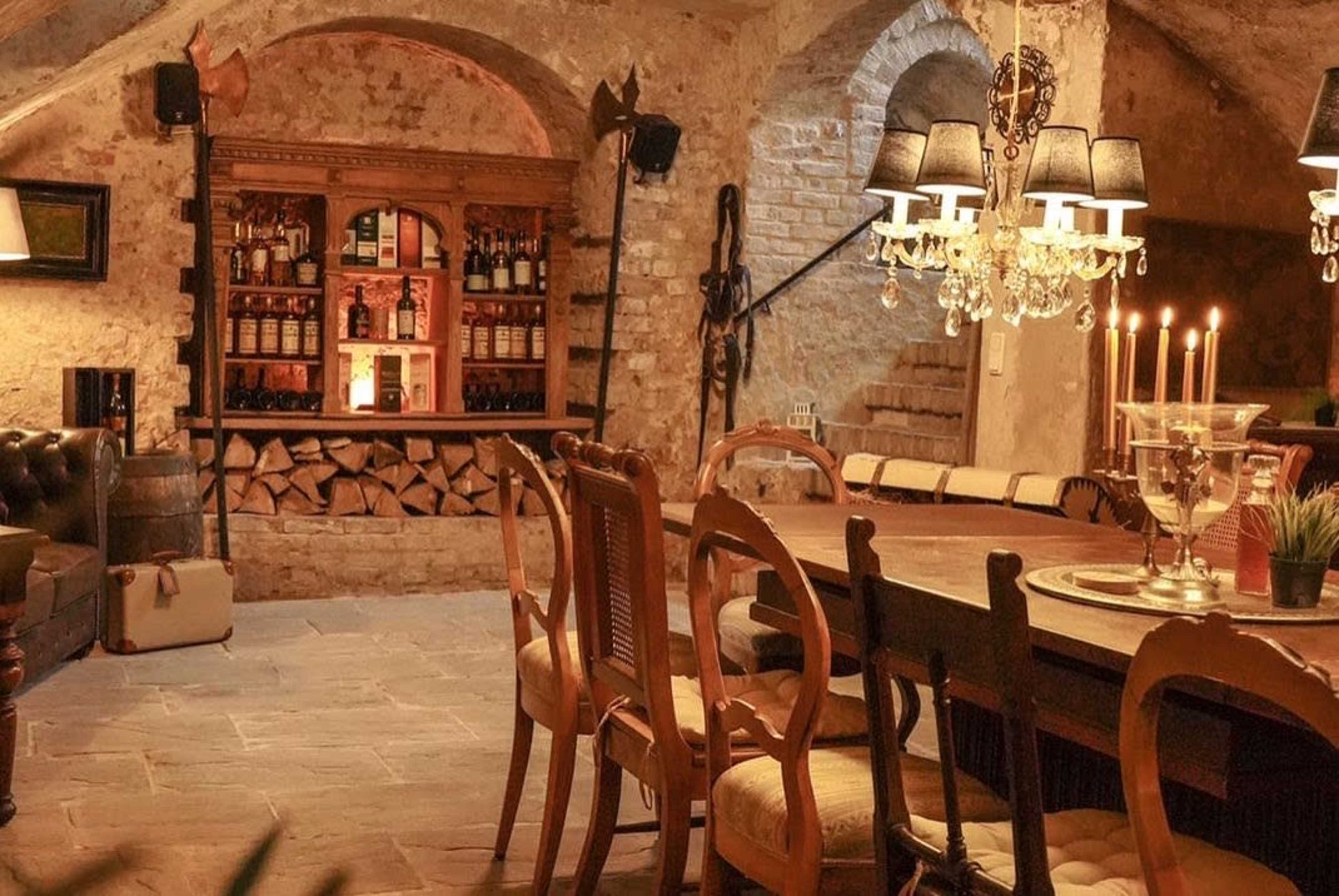 A rustic dining area featuring a large wooden table, surrounding chairs, a brick wall, and a bar with various bottles behind it.