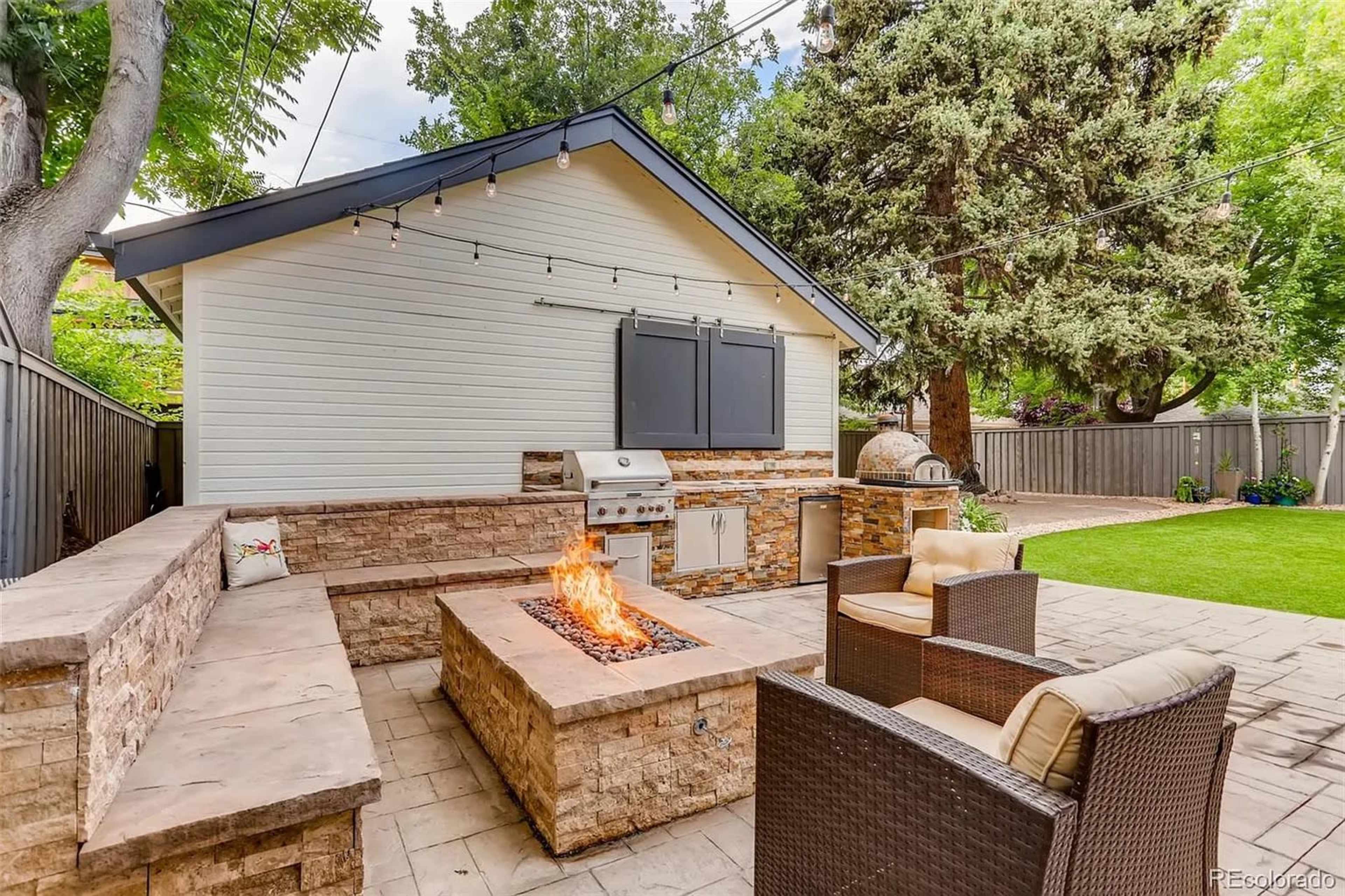 The image shows a backyard patio with a stone fire pit, outdoor seating, a built-in grill, and an adjacent tree-lined area.