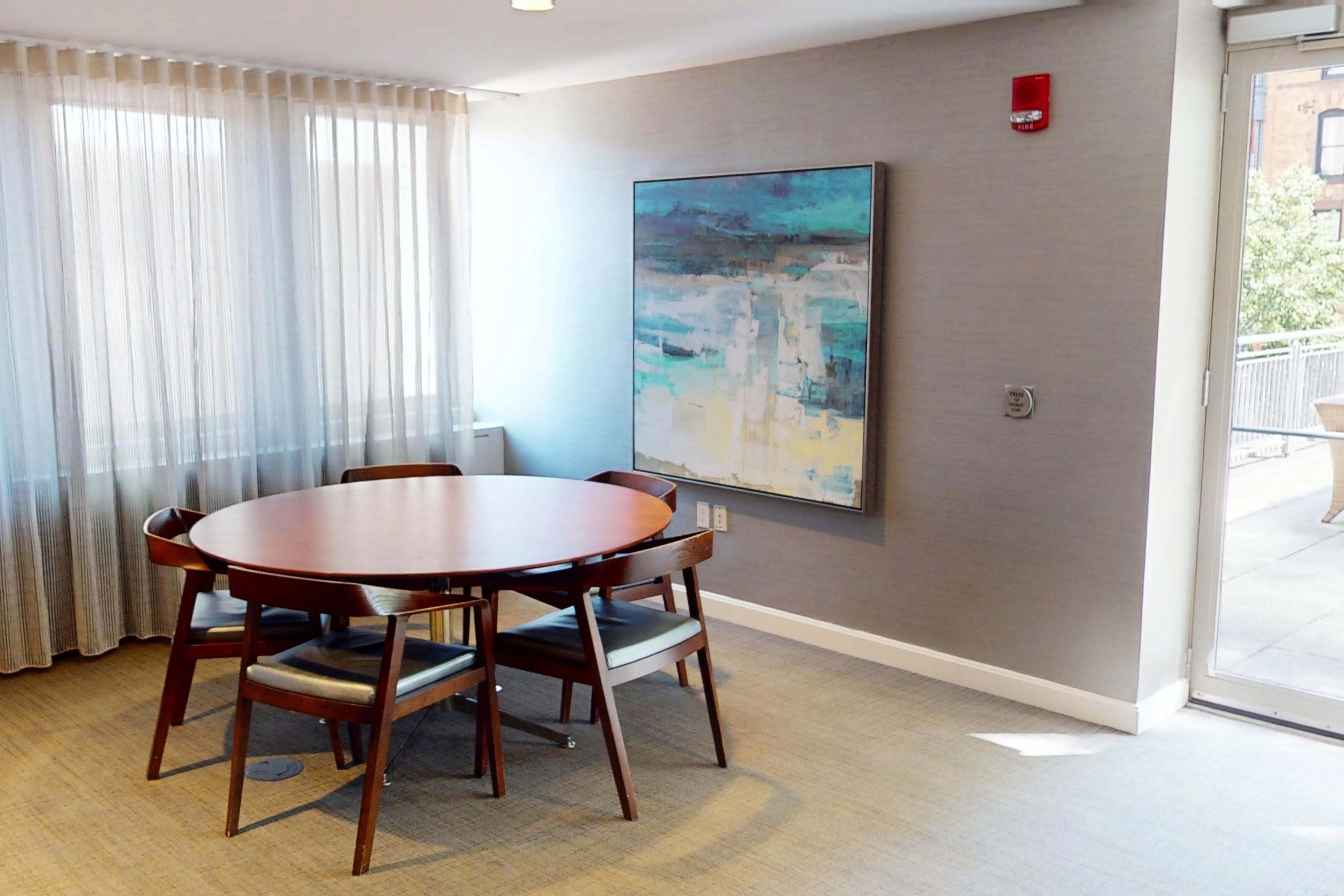 A round wooden table with four chairs is positioned in a well-lit room next to a large window, featuring an abstract painting on the wall.