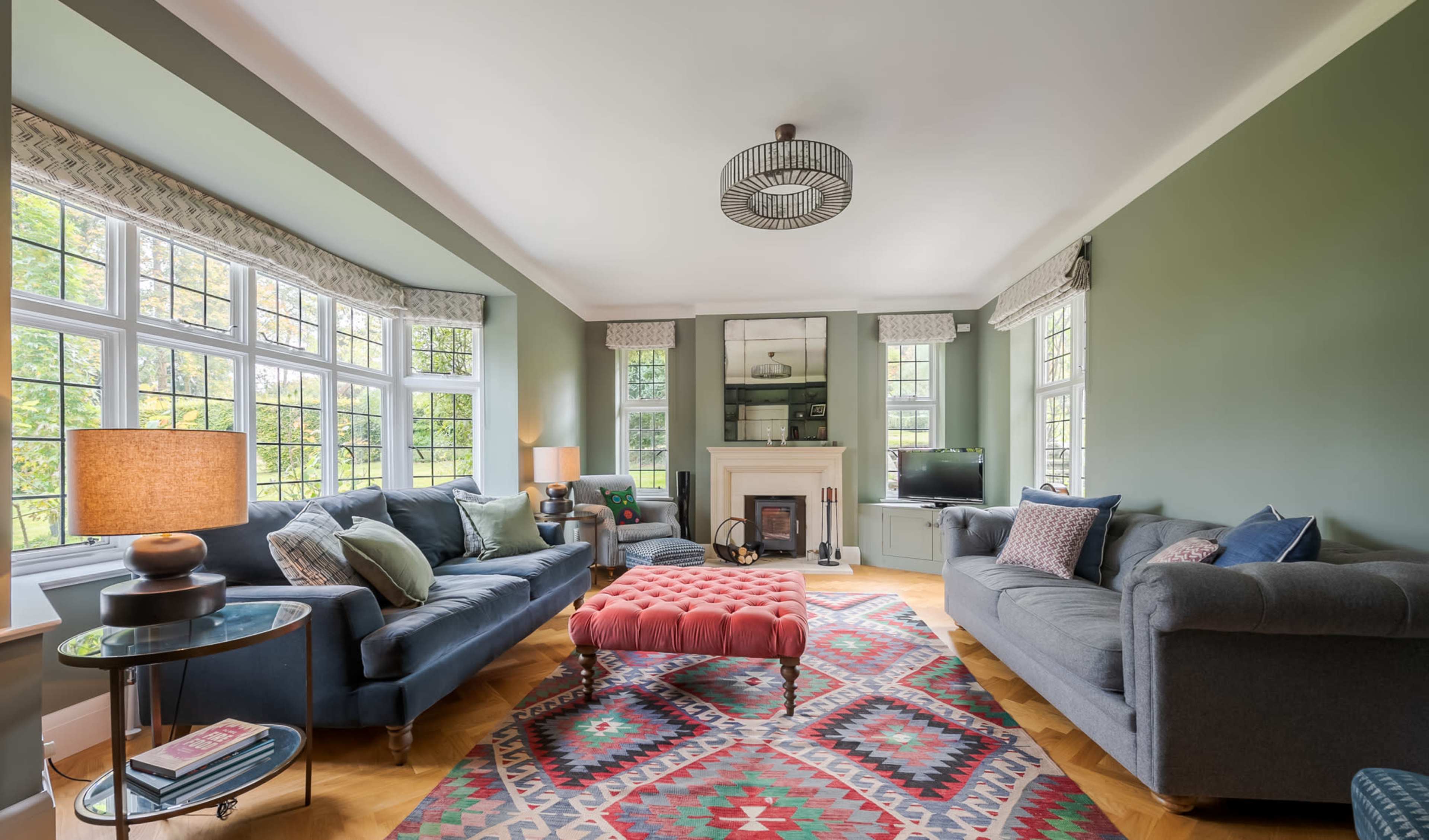 The living room features two sofas, a central ottoman, a patterned area rug, and large windows allowing natural light to fill the space.