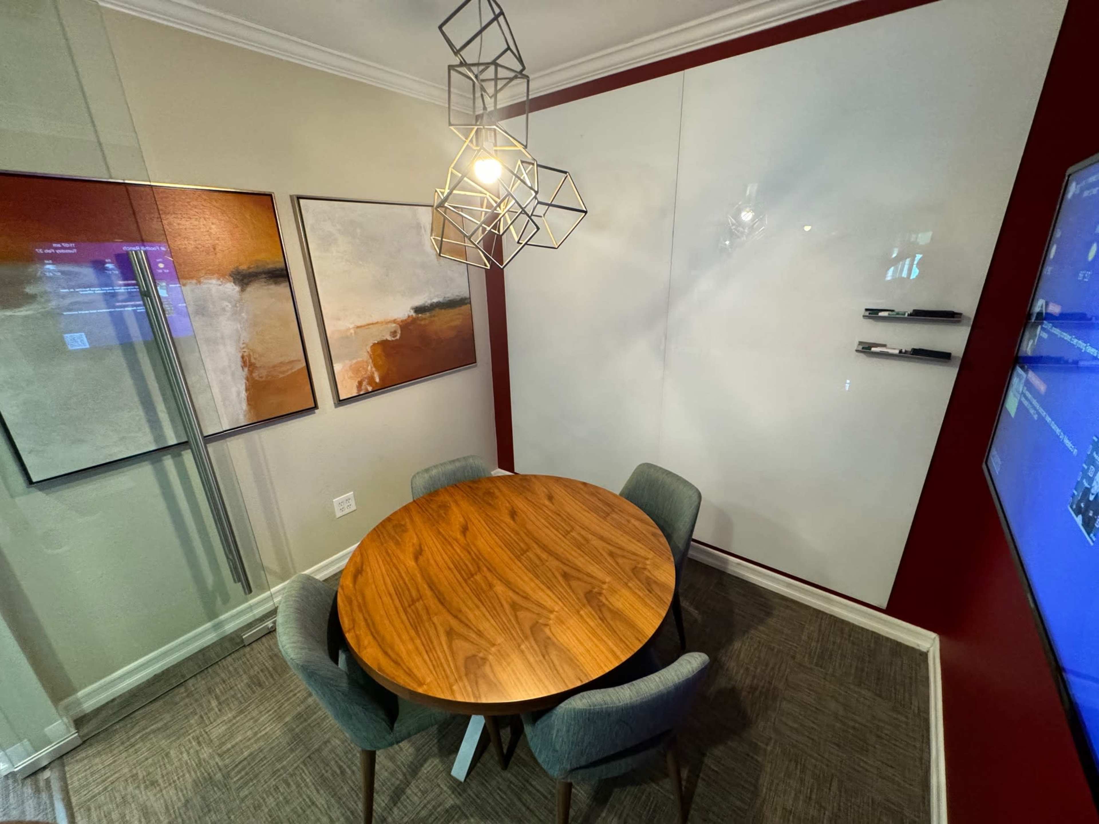 A round wooden table surrounded by four gray chairs is set in a modern meeting room featuring abstract artwork and a large screen.
