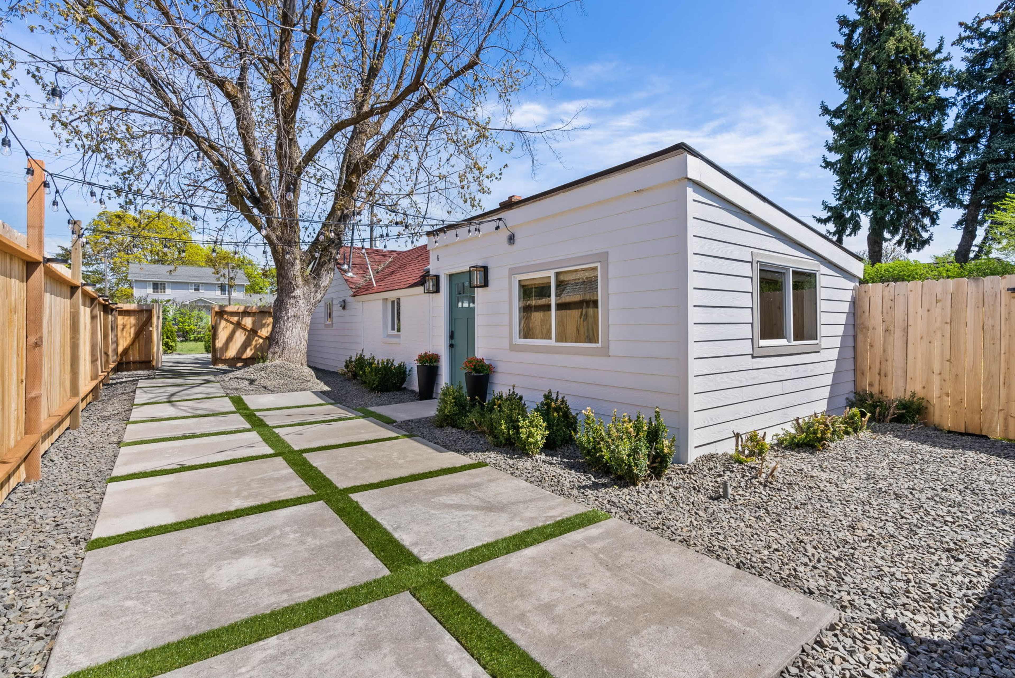 A narrow pathway lined with gravel and grass leads to a light-colored house surrounded by a wooden fence and a large tree.