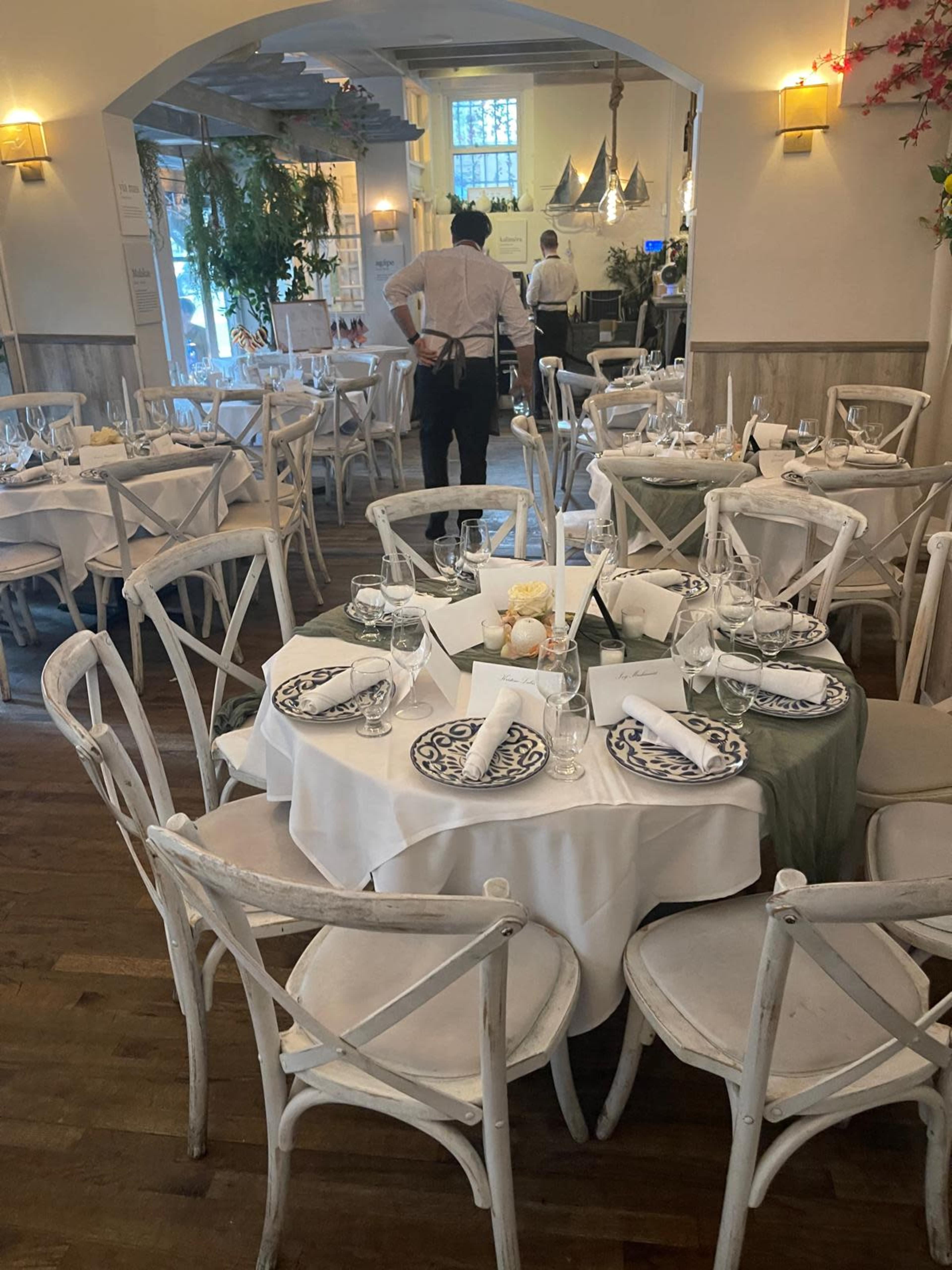 A dining area in a restaurant features several round tables set with dishes and glassware, while a waiter stands in the background.