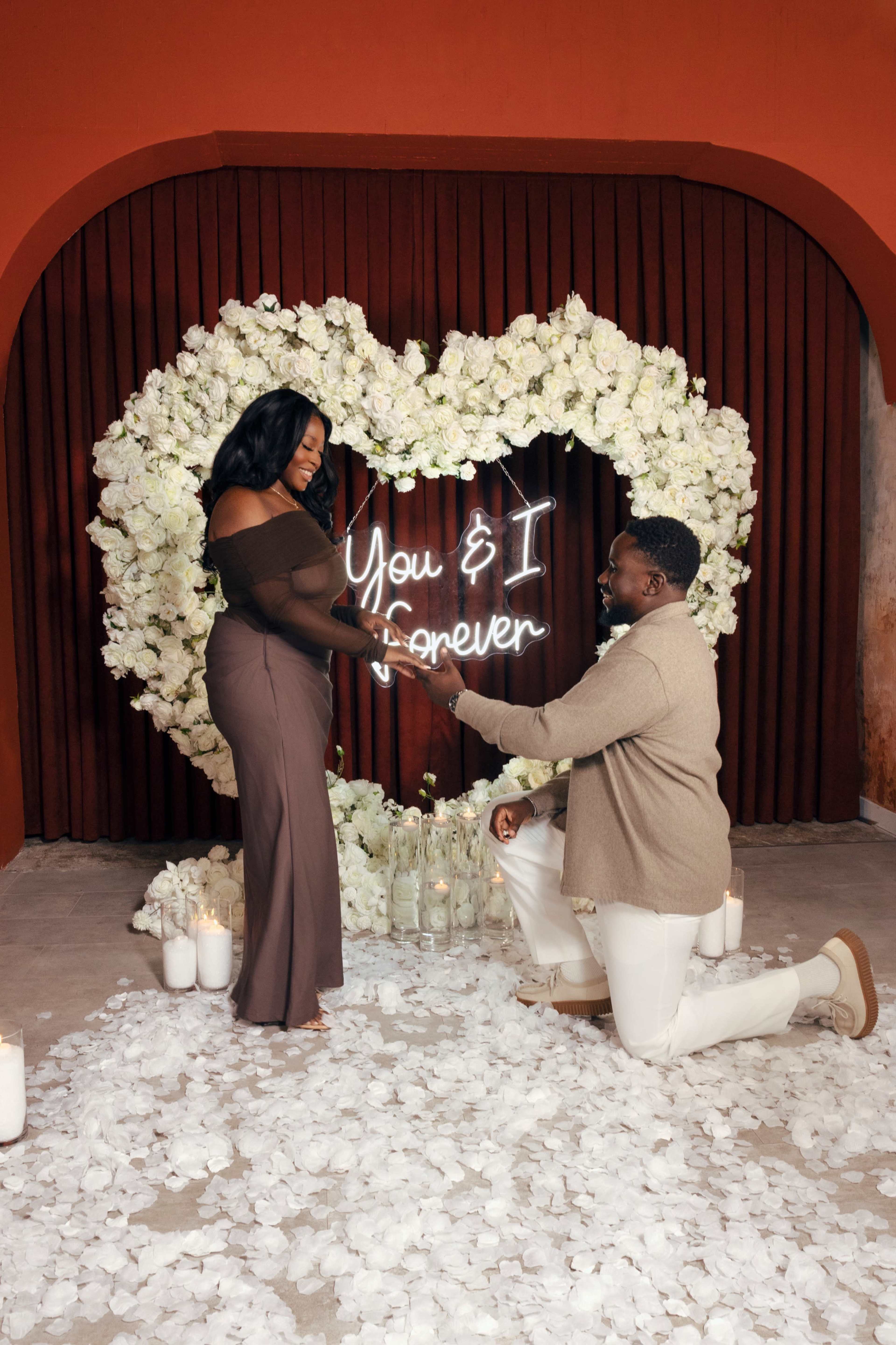 A man kneels proposing to a woman against a backdrop of a heart-shaped arrangement of white flowers, with a neon sign that reads "You & I Forever."