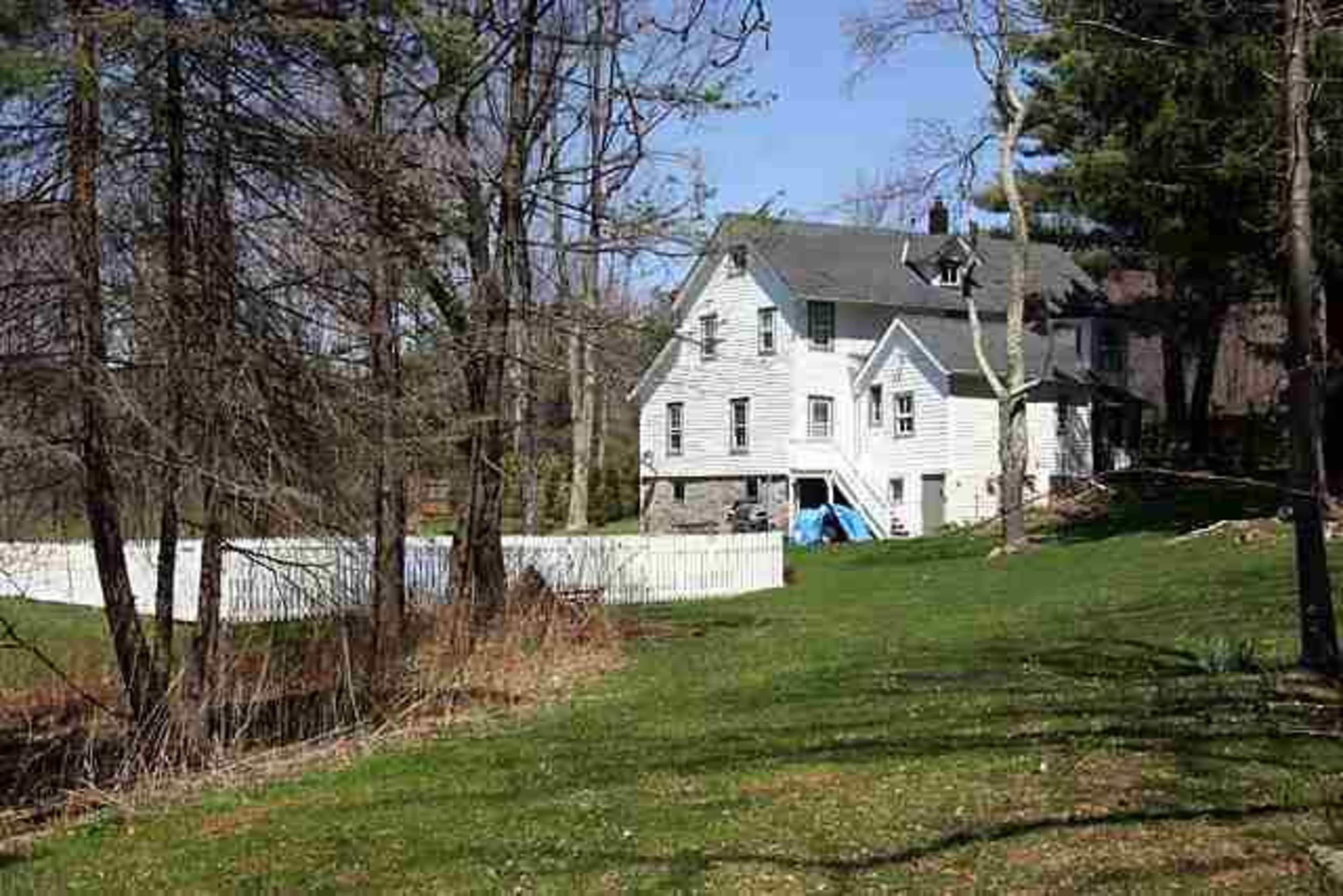 A large, two-story house is situated near a white picket fence, surrounded by trees and green grass on a sunny day.