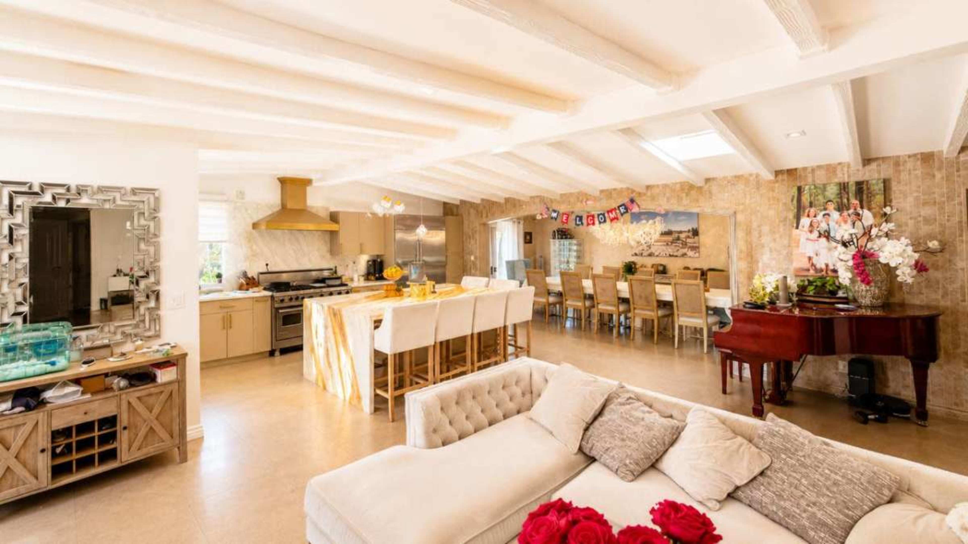 The image depicts a bright, open-concept living space featuring a large kitchen area, a dining table with multiple chairs, a piano, and a comfortable sofa adorned with decorative pillows.