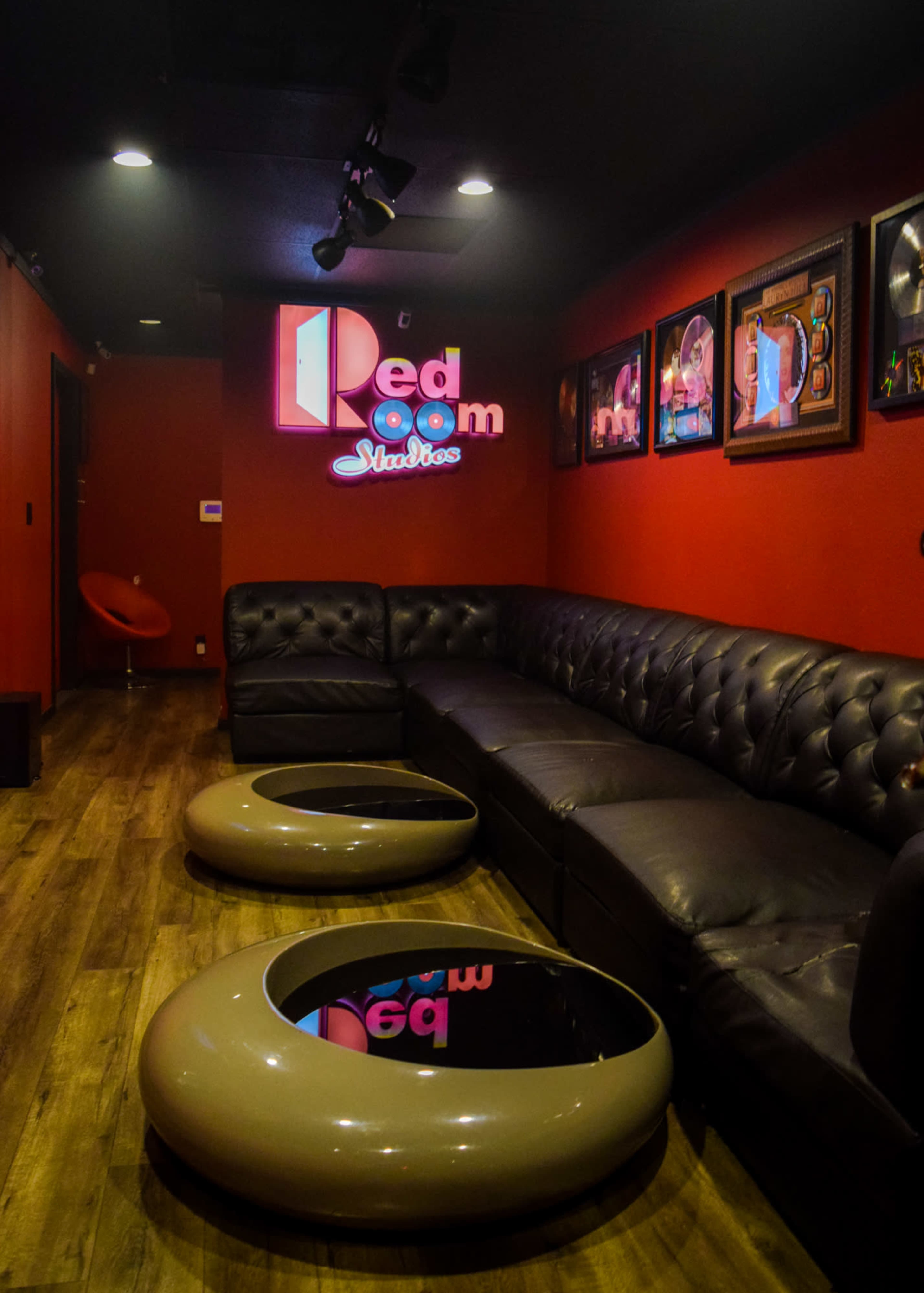 The image shows a dimly lit room featuring a black leather sectional sofa, two round coffee tables with glass tops, and colorful framed artwork on the red walls.