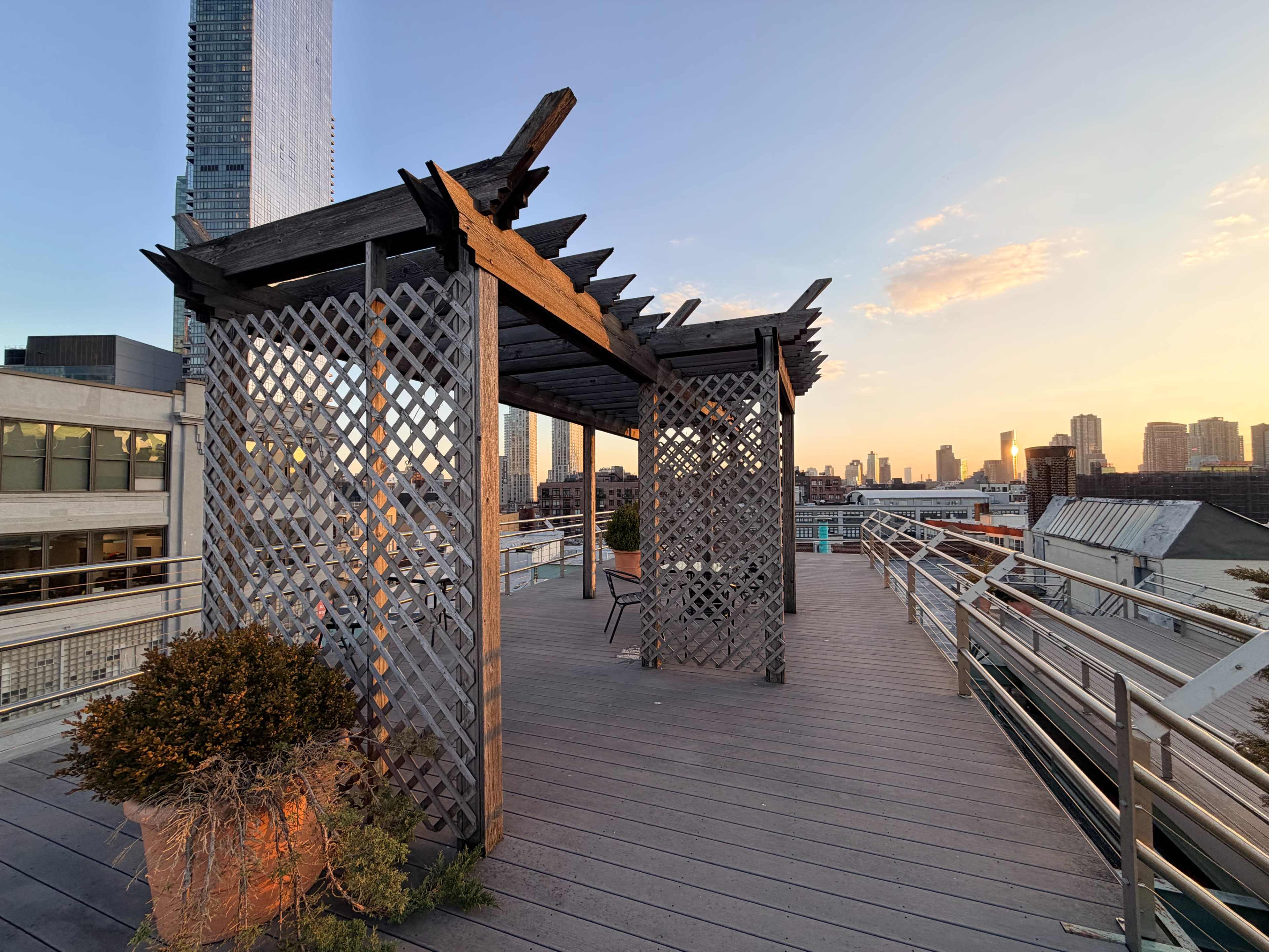 A wooden pergola stands on a rooftop terrace overlooking a city skyline at sunset.