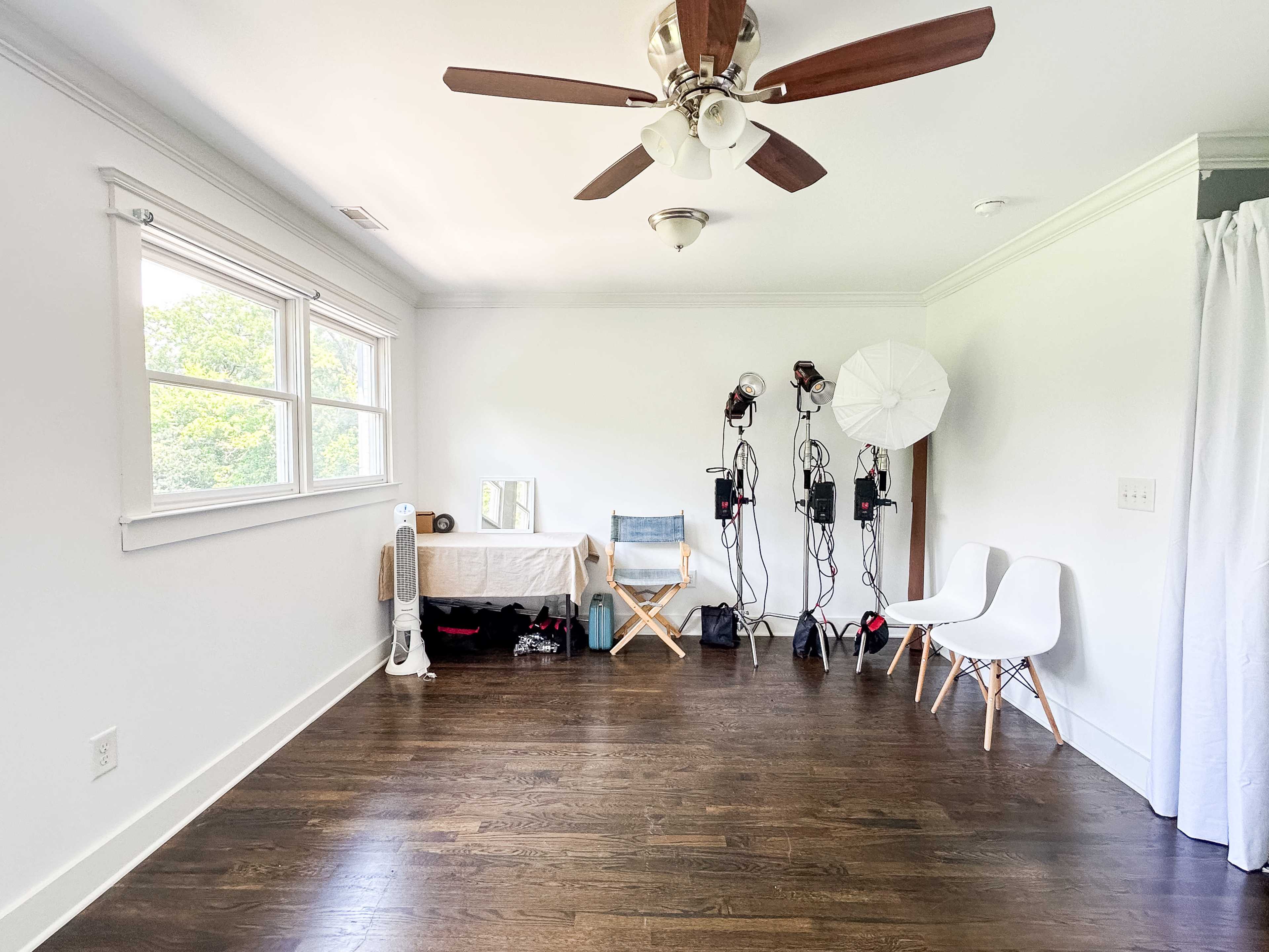 The image shows a bright, empty room with a wooden floor, a ceiling fan, and various photography equipment set up against one wall.