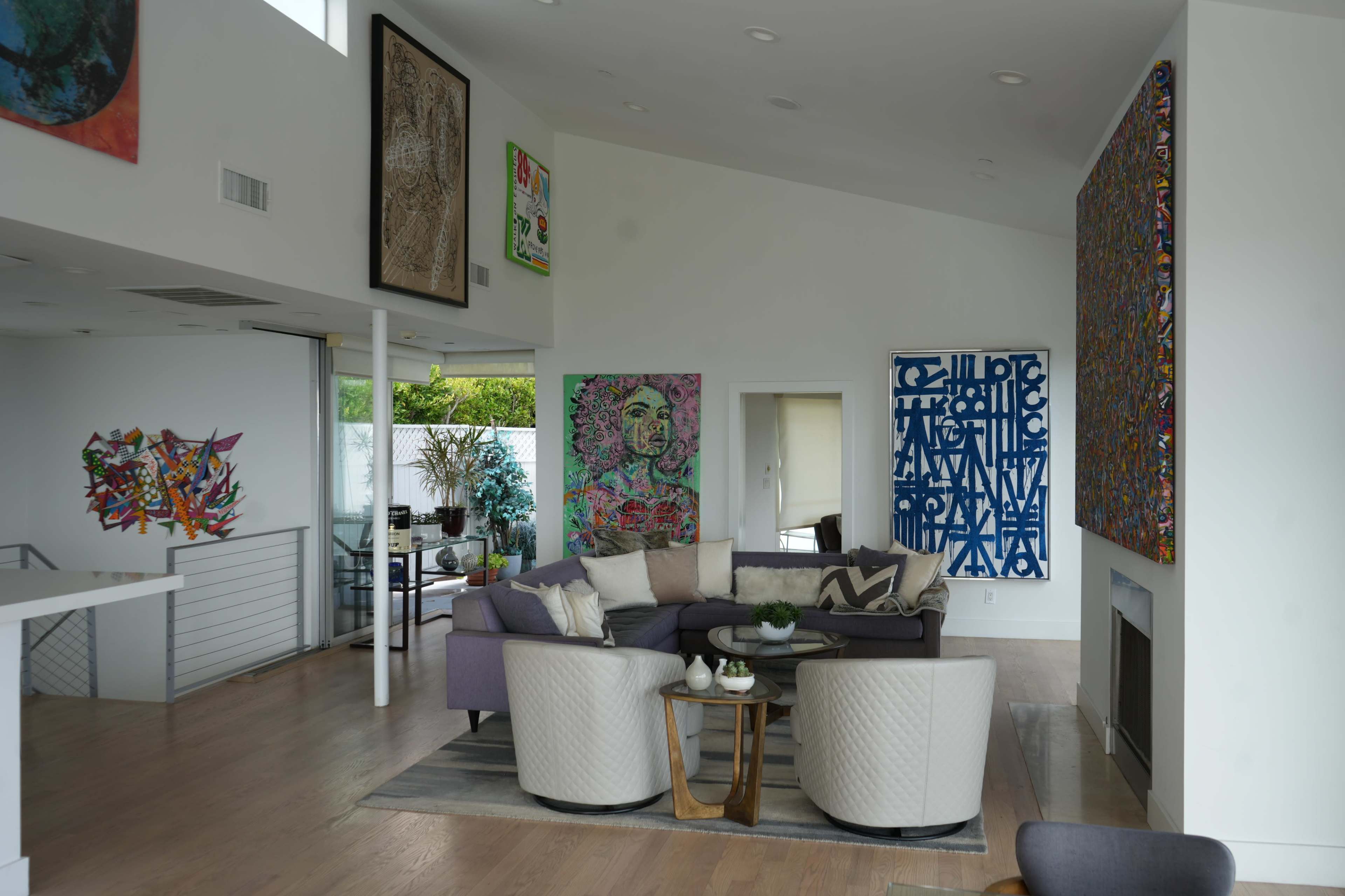 The image shows a modern living room featuring a purple sectional sofa, two light gray armchairs, and several colorful artworks displayed on the walls.