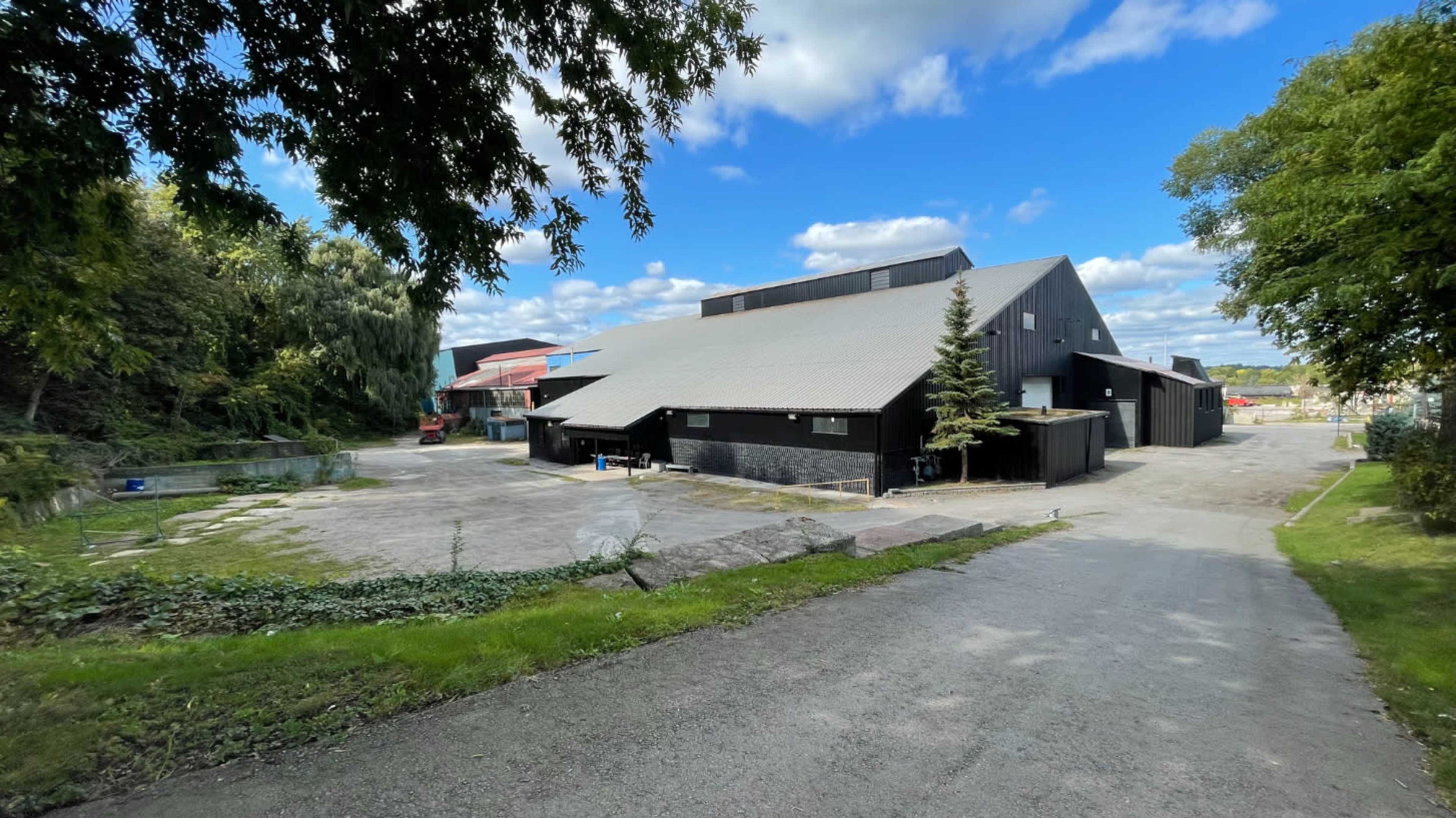 A large black building with a sloped roof is situated at the end of a gravel drive, surrounded by greenery and blue skies.