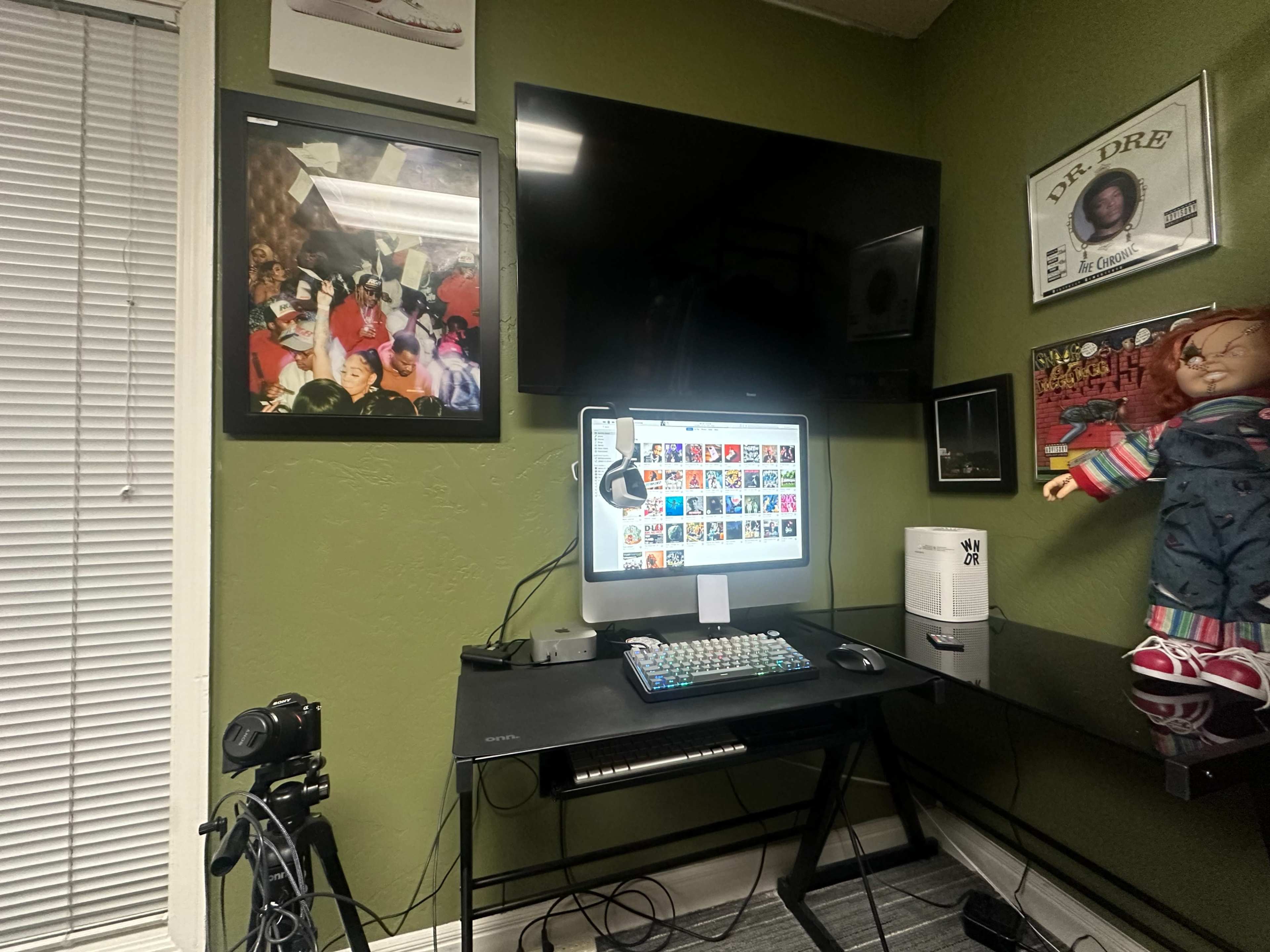 The image shows a workspace featuring a computer monitor displaying colorful thumbnails, a black desk, a camera on the left, a television on the wall, and various framed pictures and decorations.