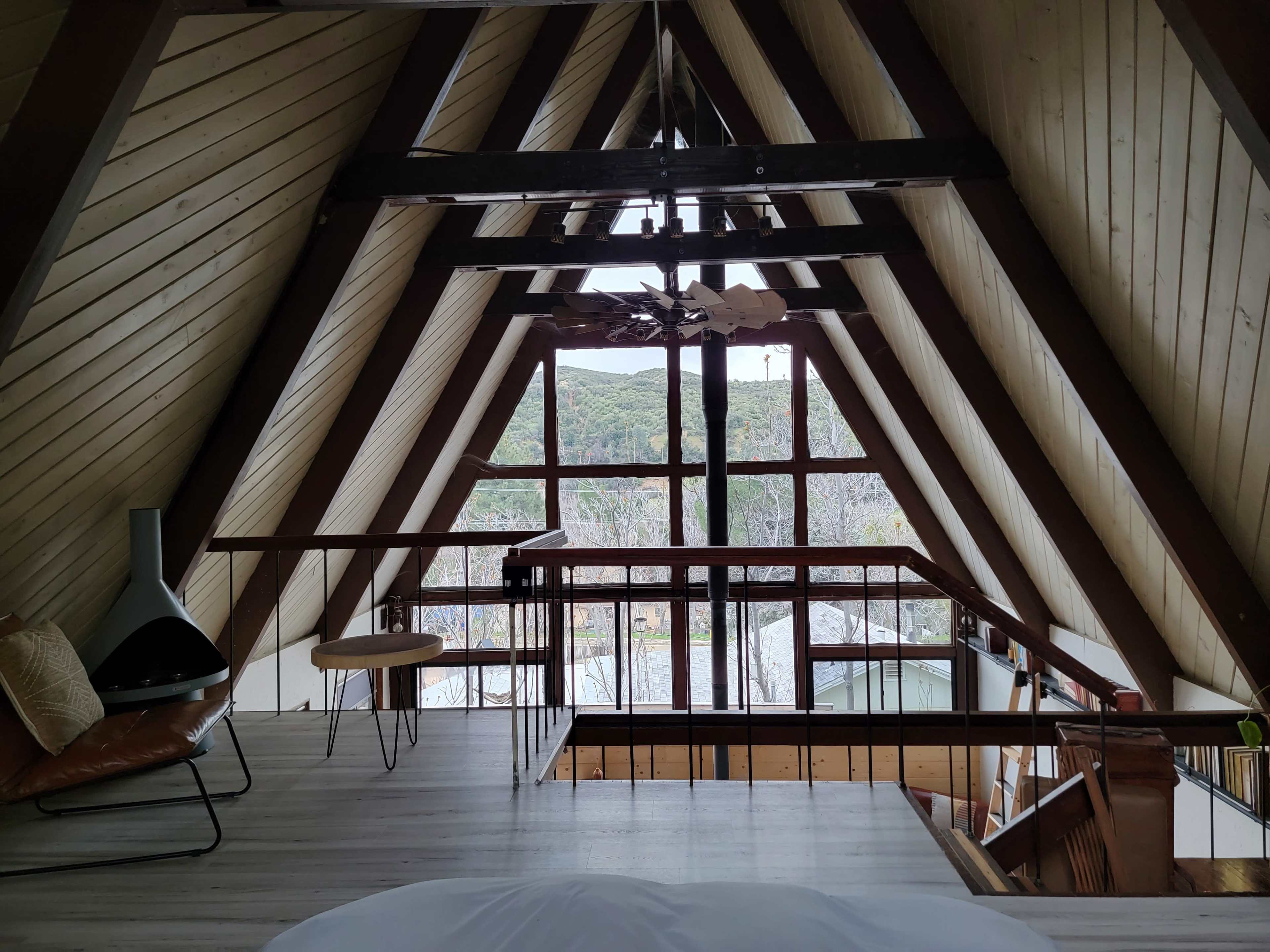 The image shows an A-frame interior with wooden beams, a balcony overlooking a landscape, and a minimalist seating area.