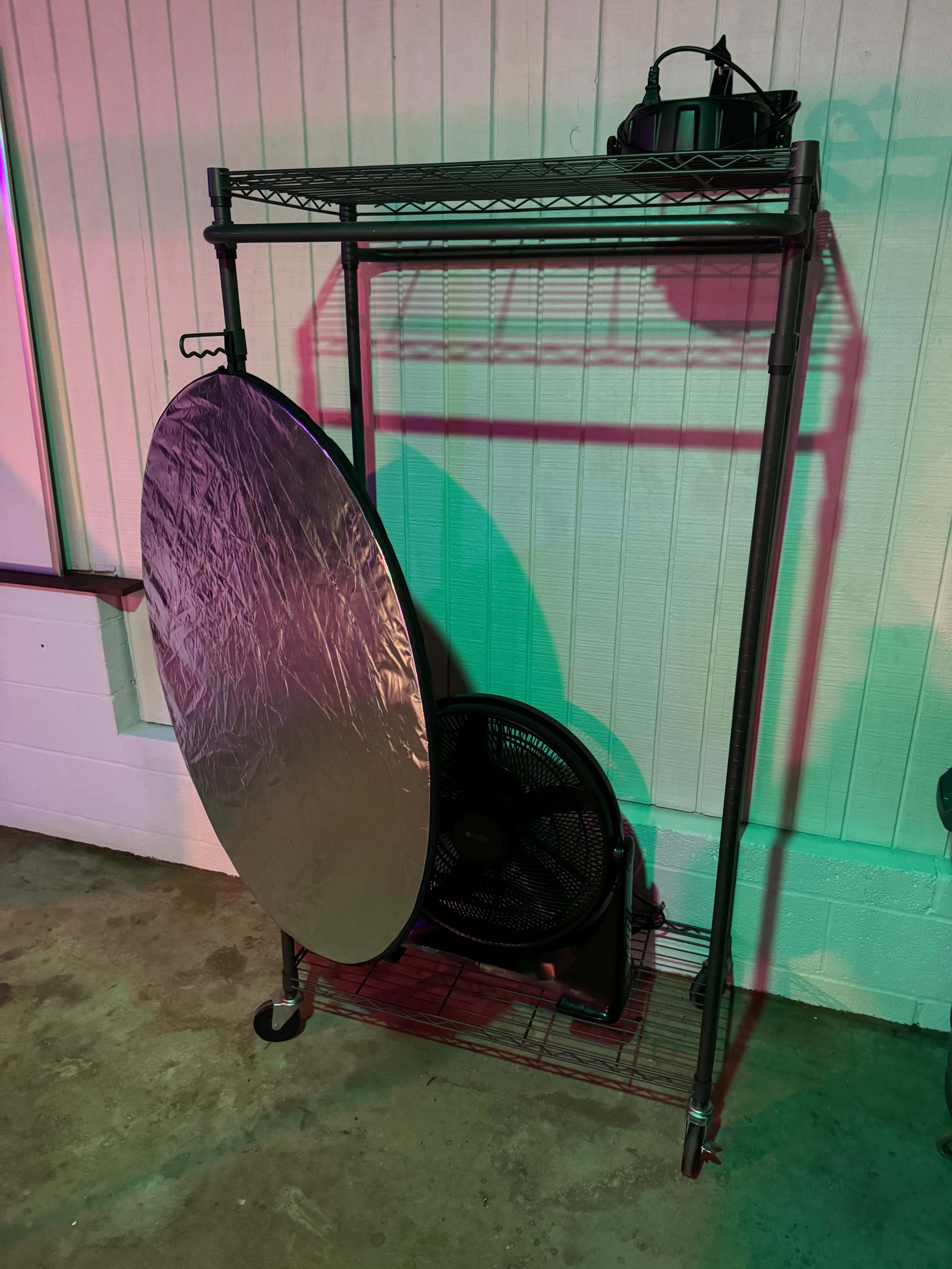 A metal shelving unit holds a large circular reflector and a standing fan in a dimly lit space.
