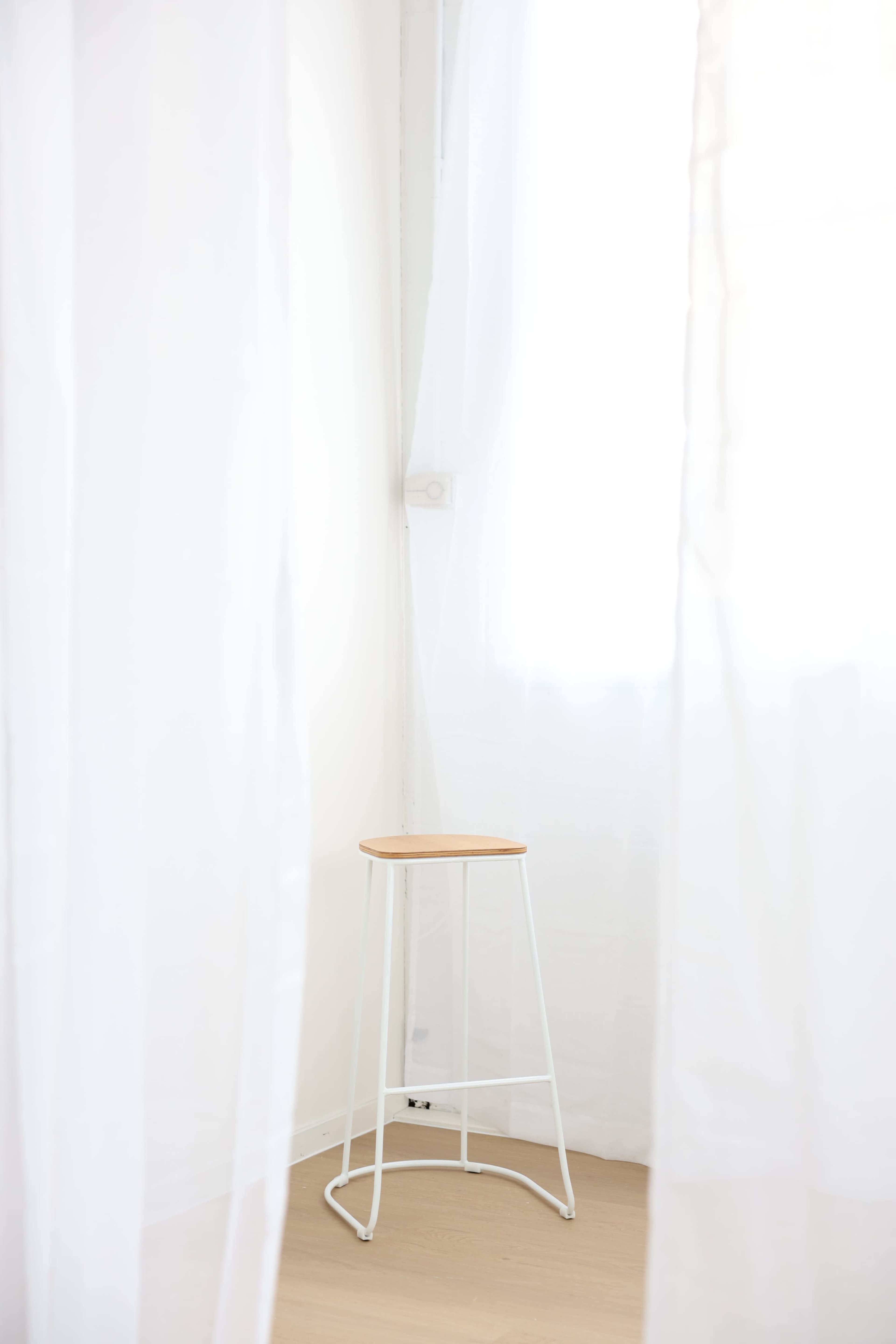 A wooden stool sits in the corner of a bright room, partially obscured by sheer white curtains.
