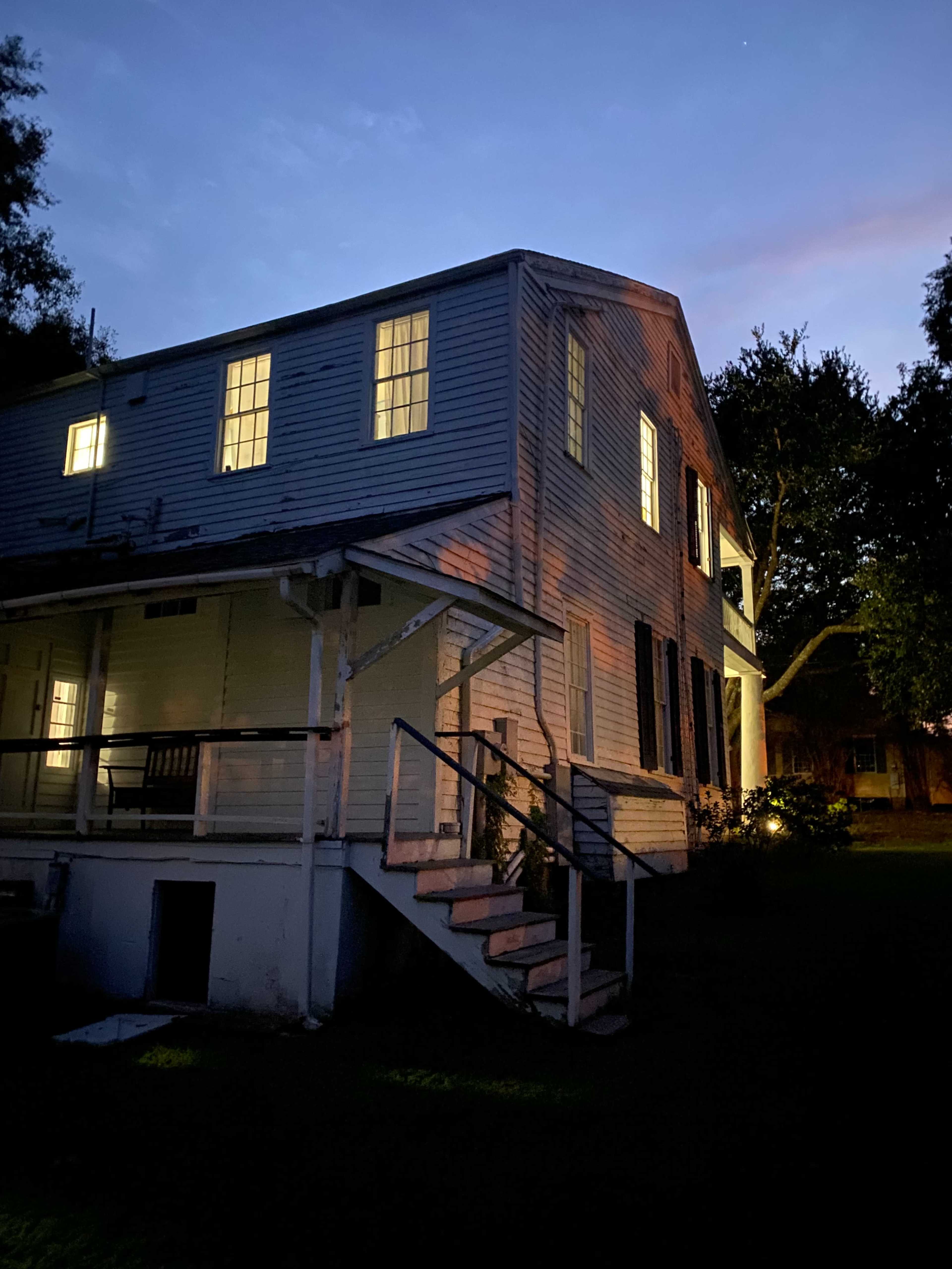 A two-story wooden house with illuminated windows sits under a twilight sky, showcasing its weathered exterior and a staircase leading to the porch.