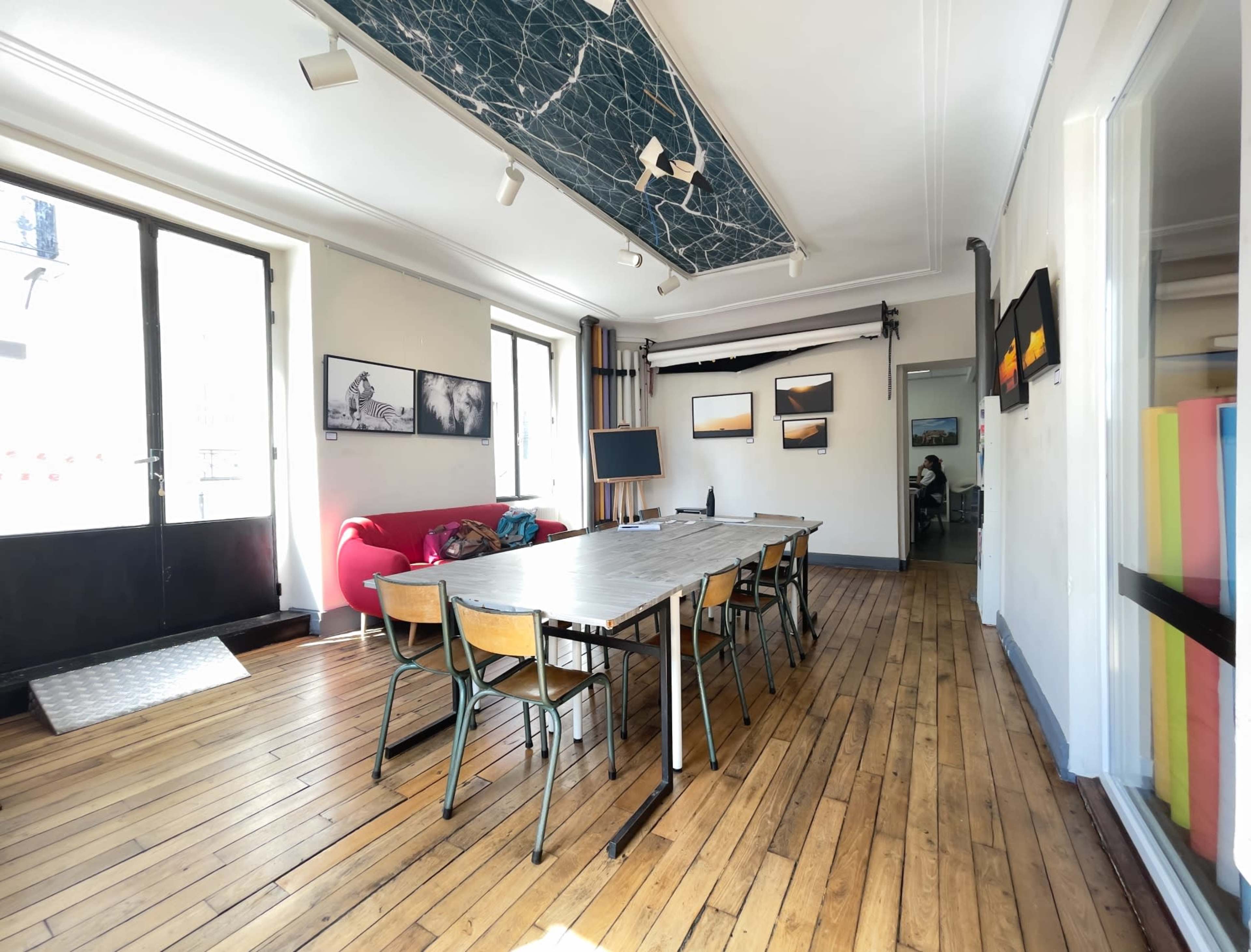 A bright room with wooden floors, featuring a long table surrounded by chairs, a red couch against one wall, and artwork displayed on the walls.