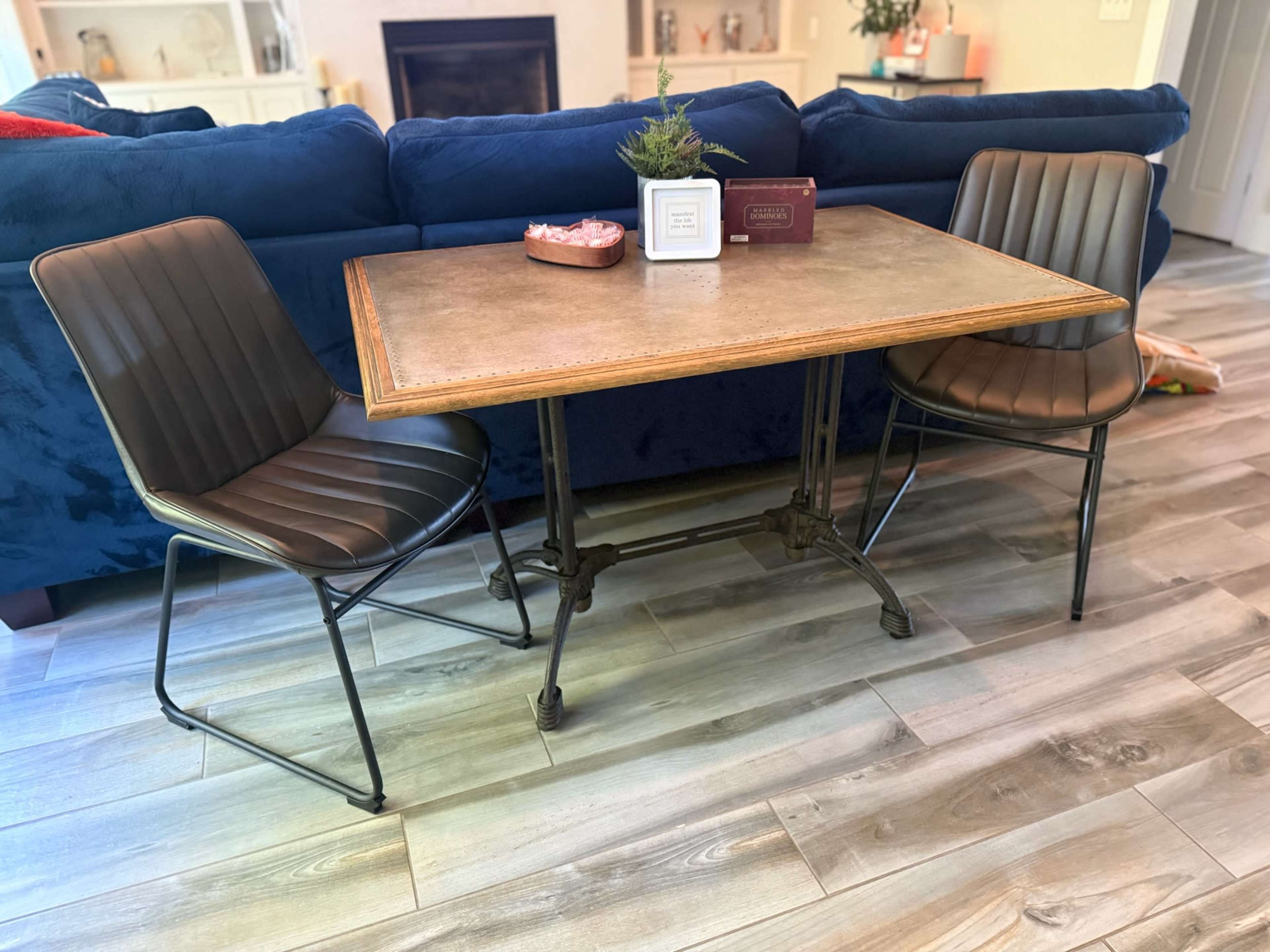 A rectangular dining table with a wooden top and metal legs is set between two leather chairs in a room with wooden flooring and a blue couch in the background.