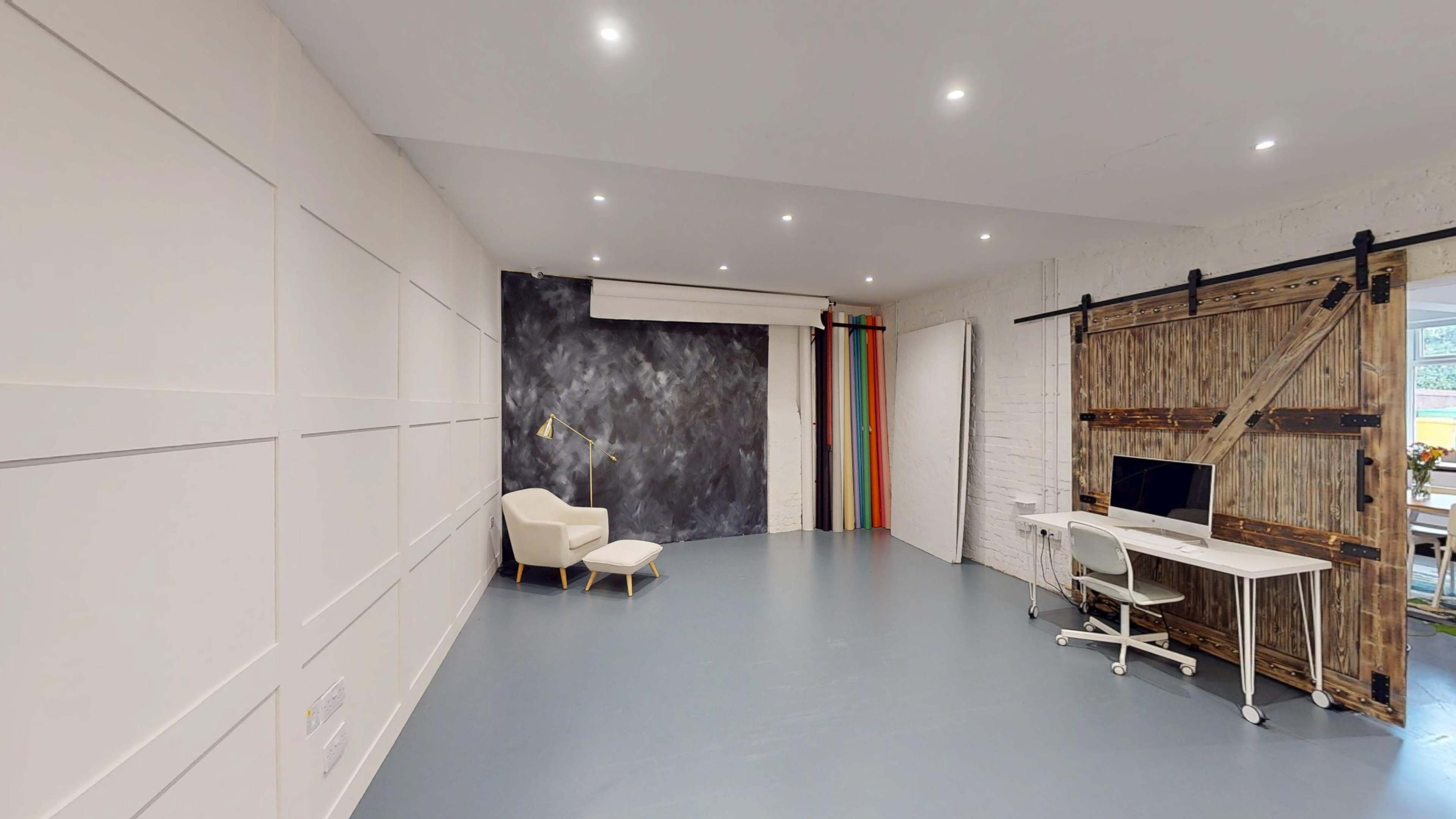 The image shows a spacious, well-lit room featuring a white wall with decorative paneling, a painted accent wall, a modern chair, and a desk with a computer.