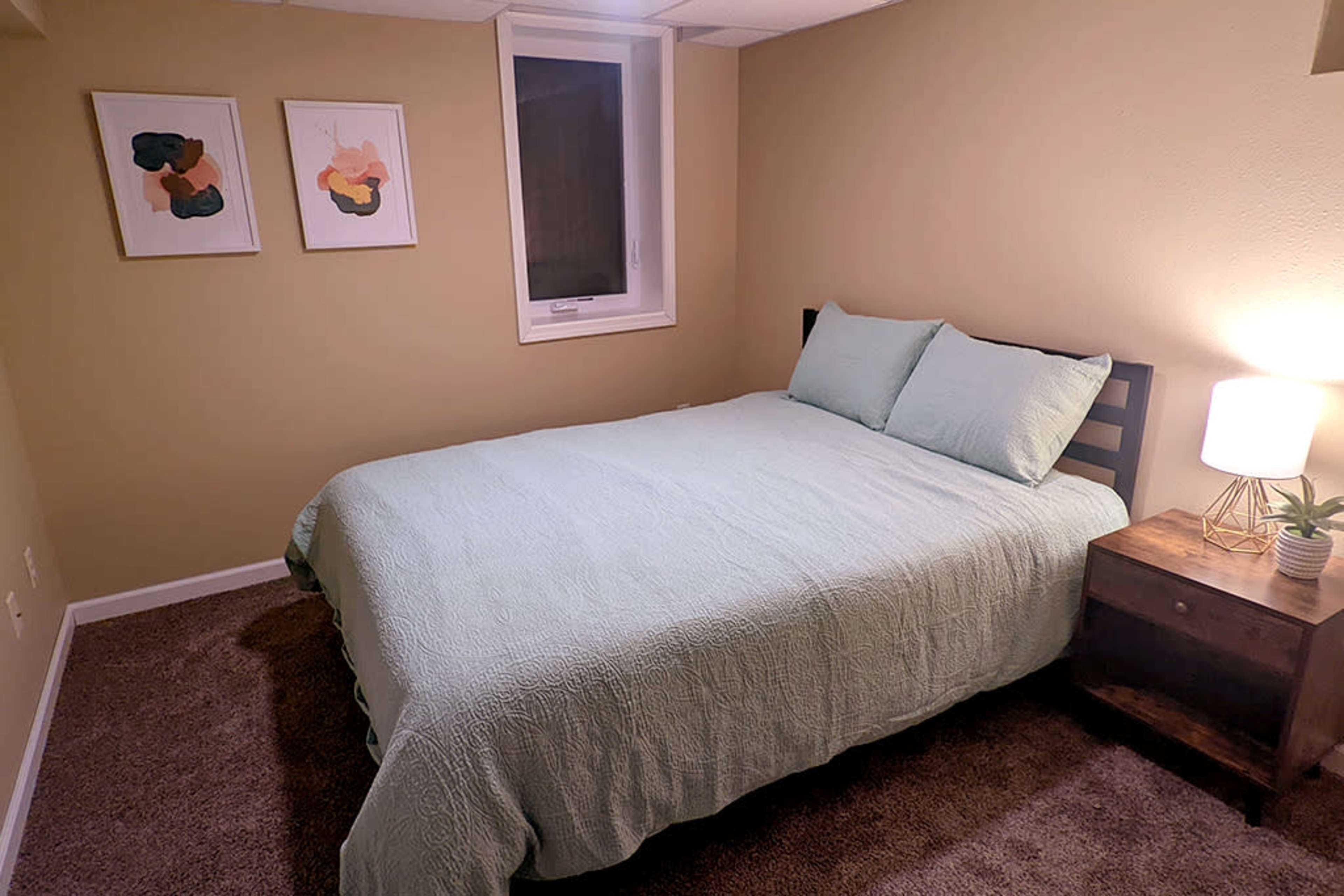 The image shows a small bedroom with a light green bedspread, a bedside table with a lamp, and two abstract art pieces hanging on the wall.