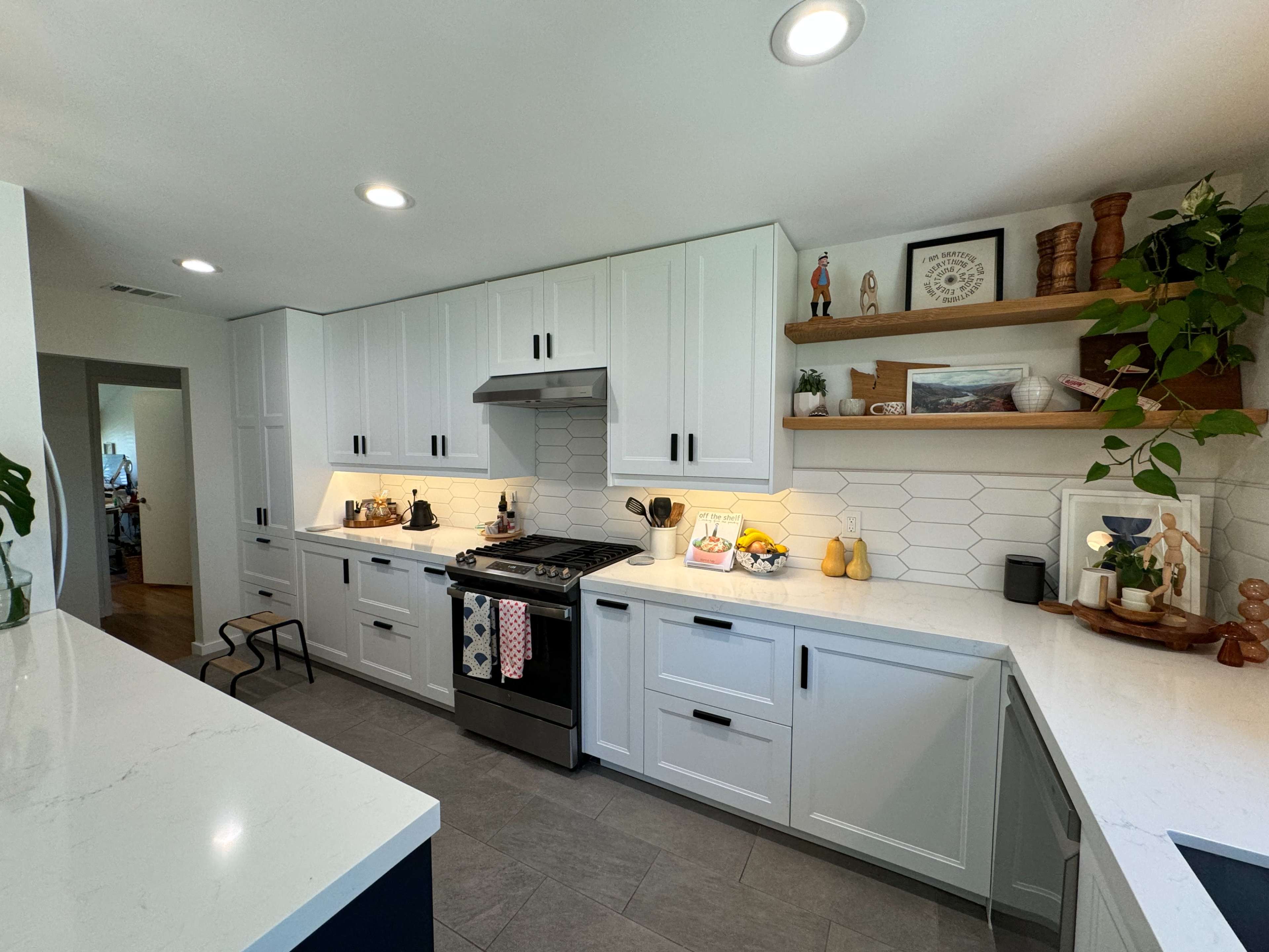 The image shows a modern kitchen with white cabinets, a stainless steel stove, and a hexagonal tile backsplash, featuring decorative items on the shelves.