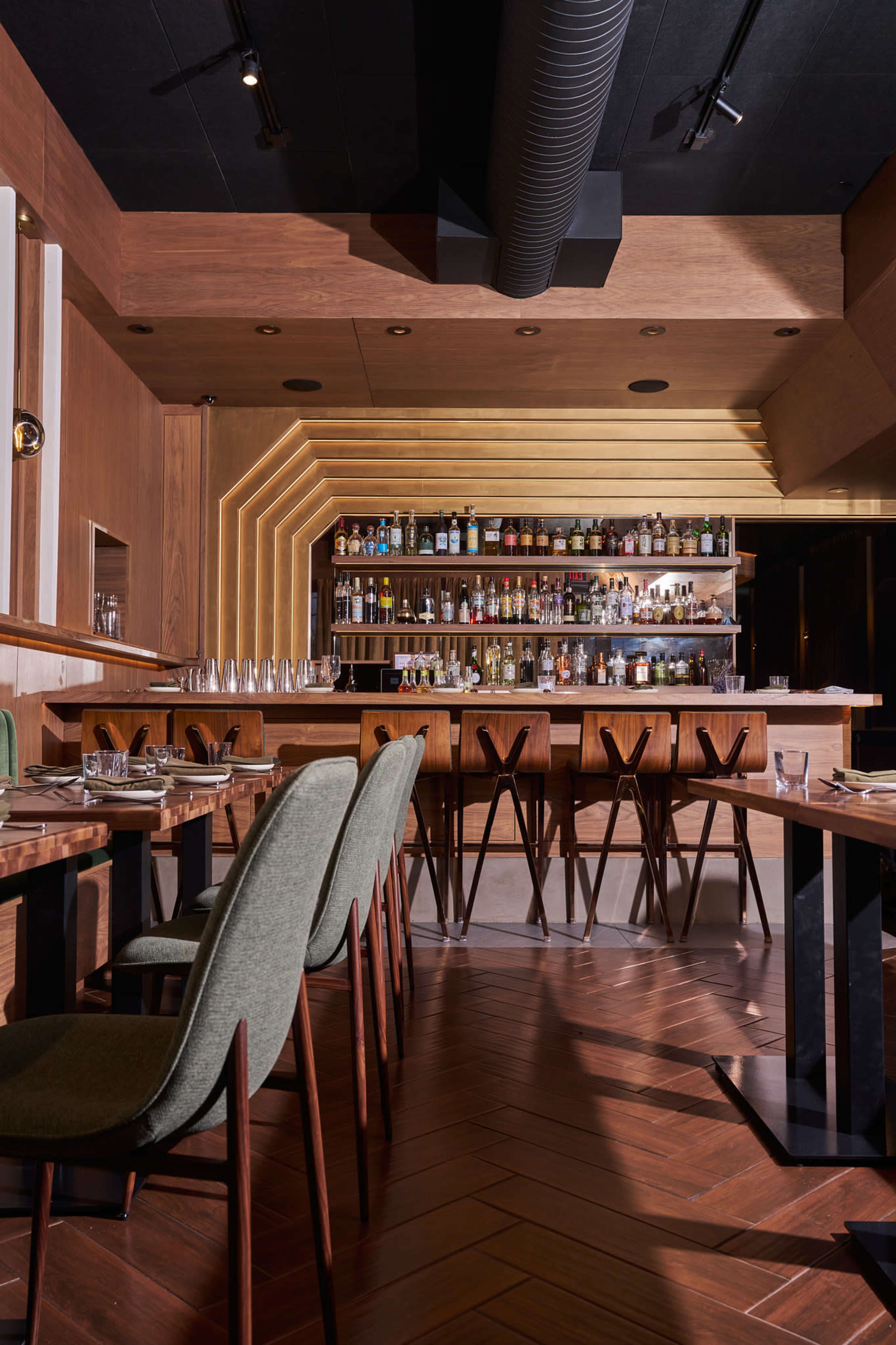 The image shows a modern restaurant interior featuring wooden walls, a well-stocked bar, and elegant seating arrangements with green chairs around tables.
