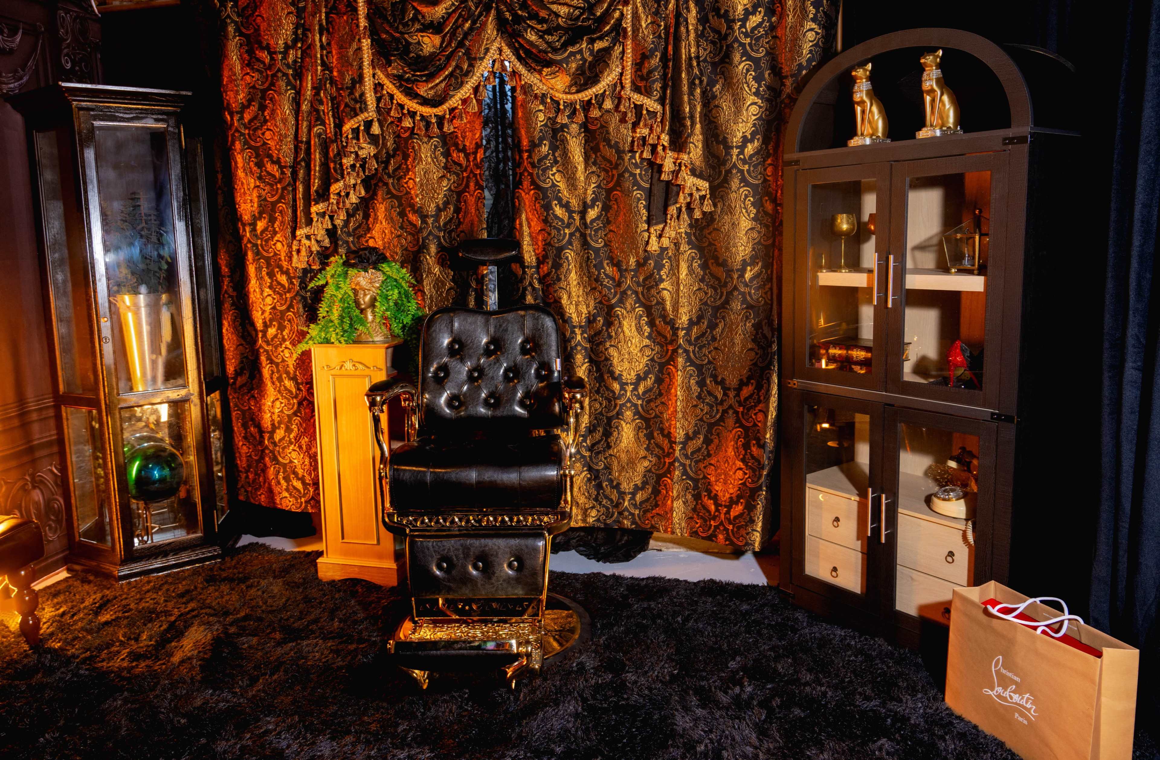The image features a vintage-style barber chair in front of ornate curtains, surrounded by decorative furniture and a display cabinet on one side.