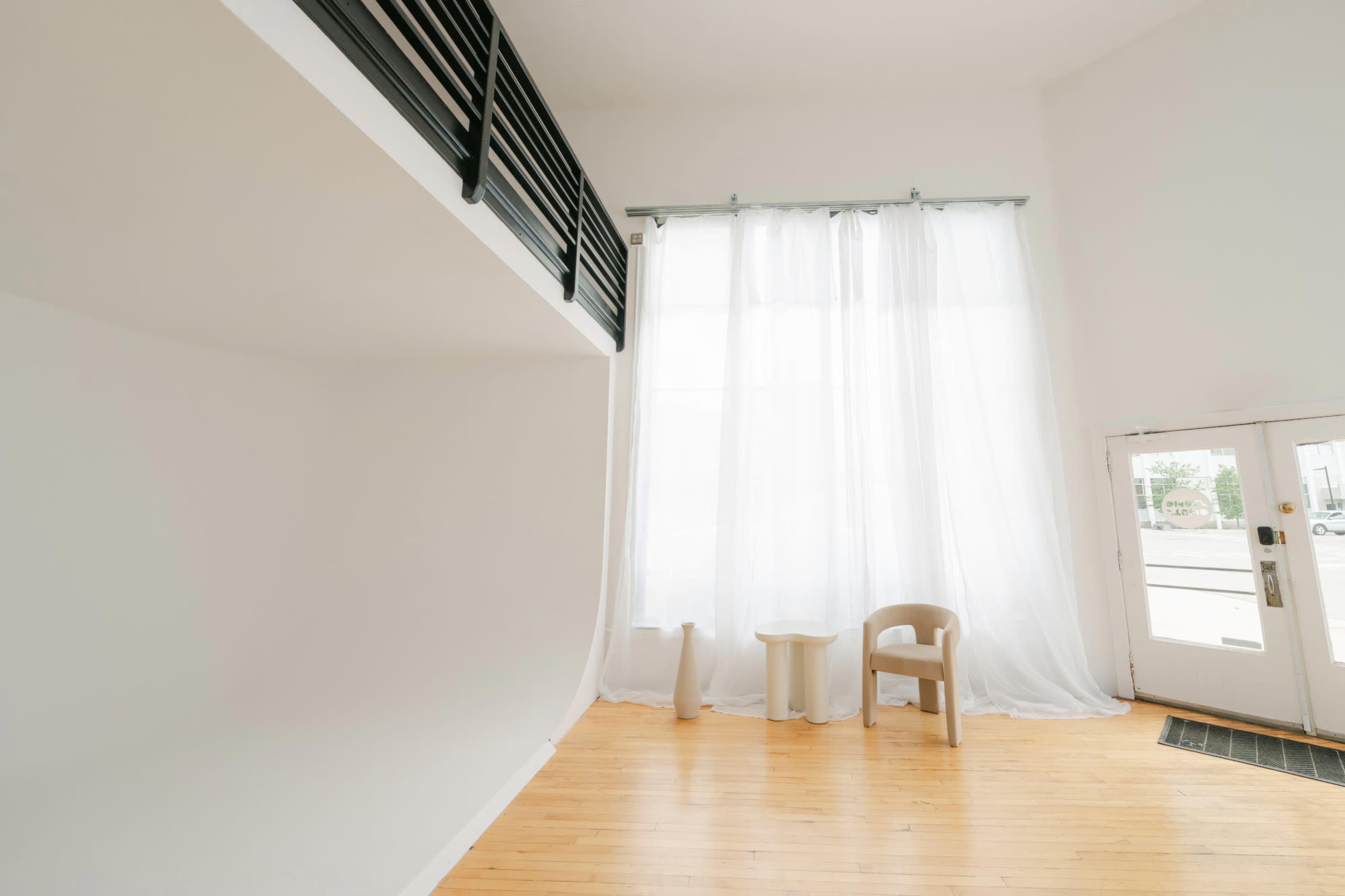 A spacious, minimalistic interior with large windows covered by sheer curtains, a small round table, and a single chair.