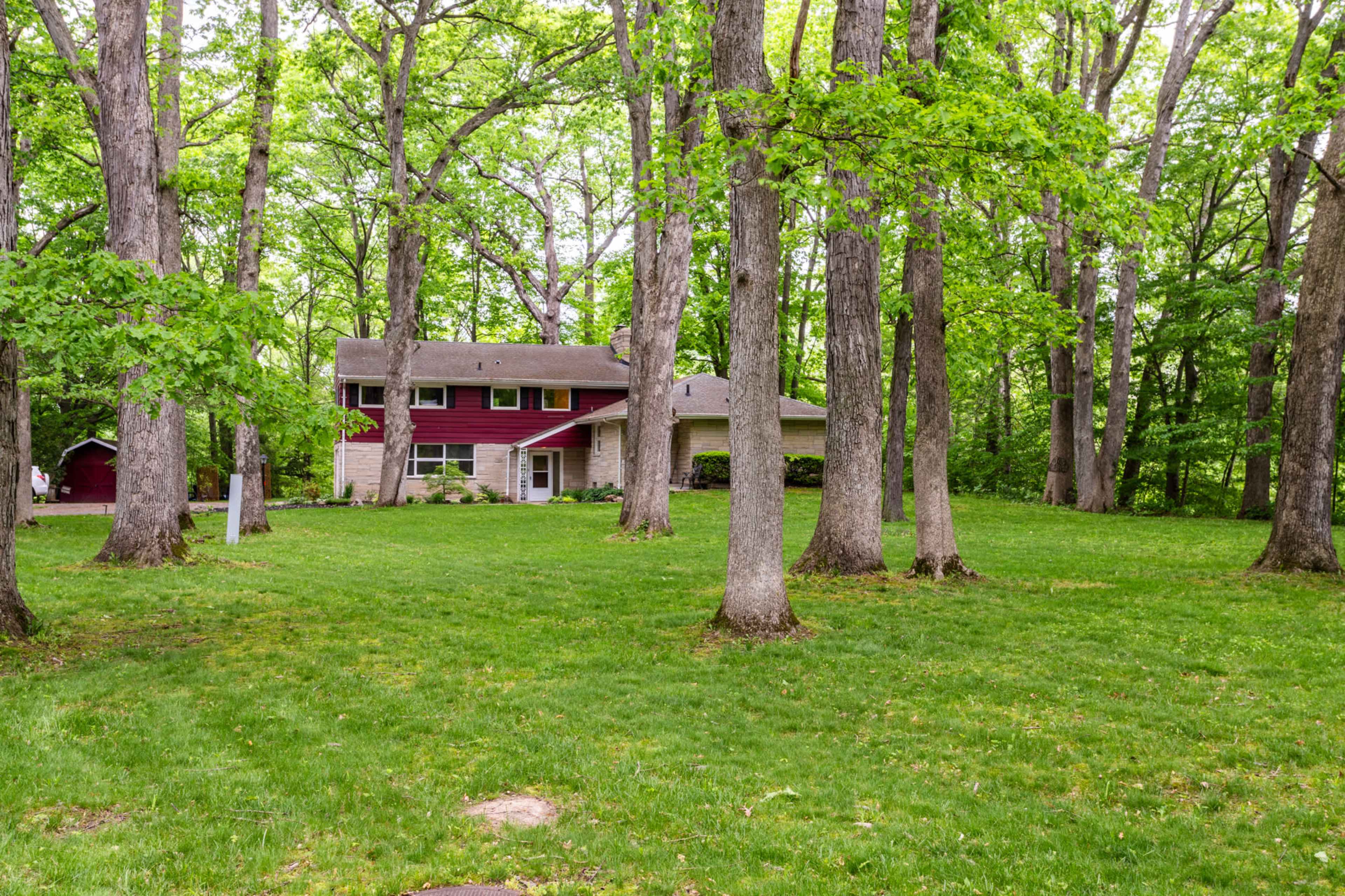 A two-story house with a red and beige facade is situated among tall trees on a grassy lot.