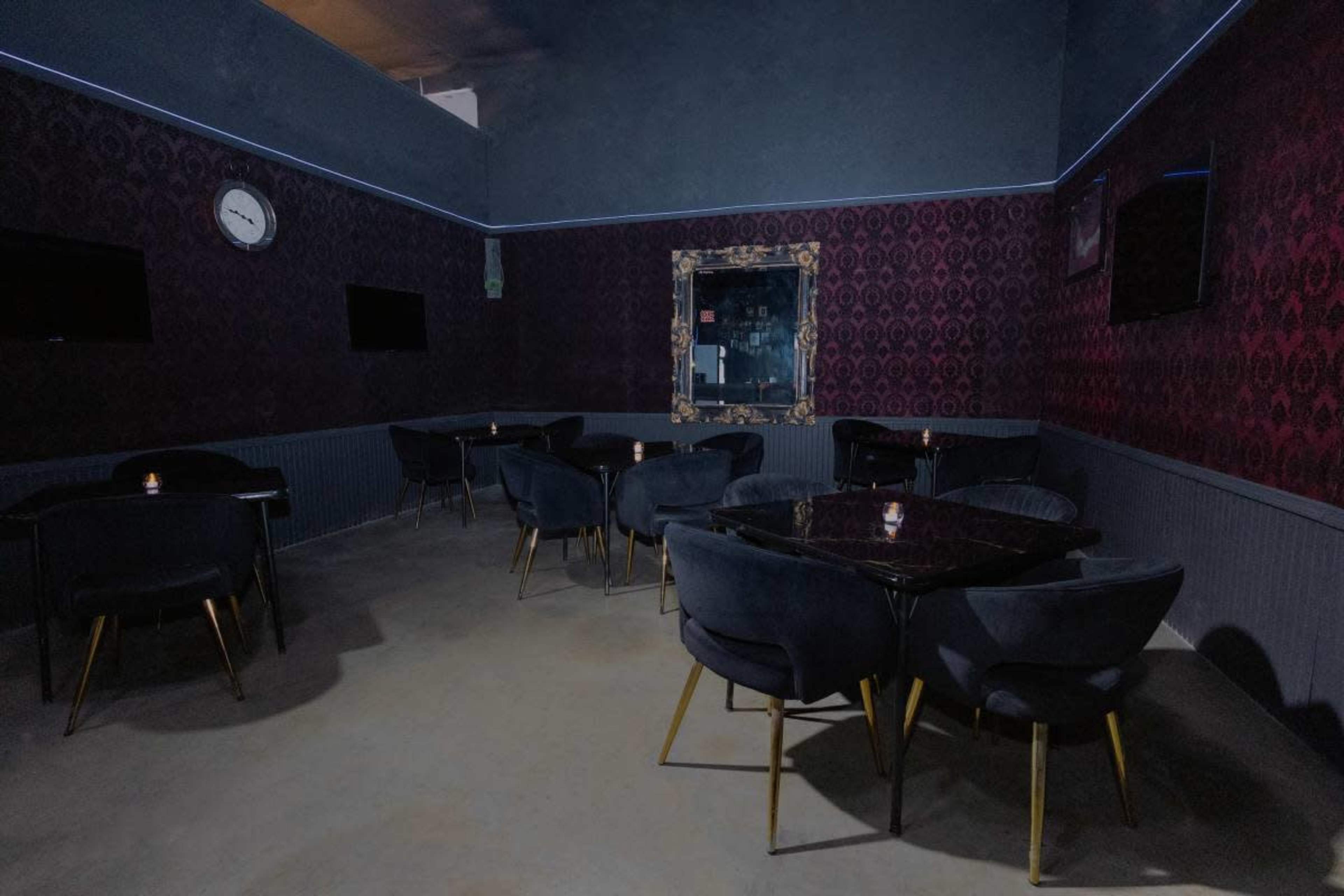 The image depicts a dimly lit room with dark patterned walls, featuring several round tables surrounded by velvet chairs.
