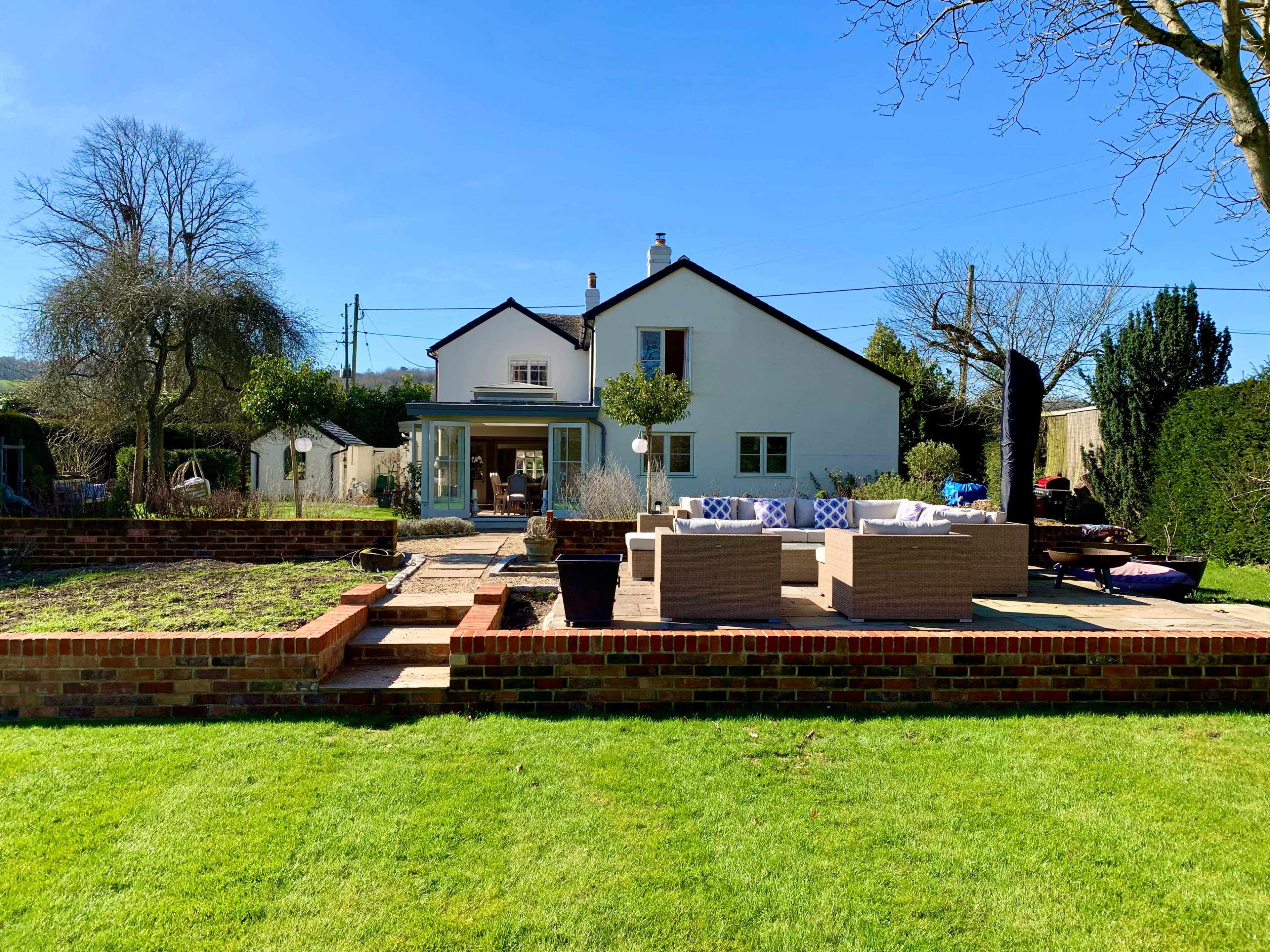 The image shows a two-story house with a patio area surrounded by green grass and a garden, under a clear blue sky.