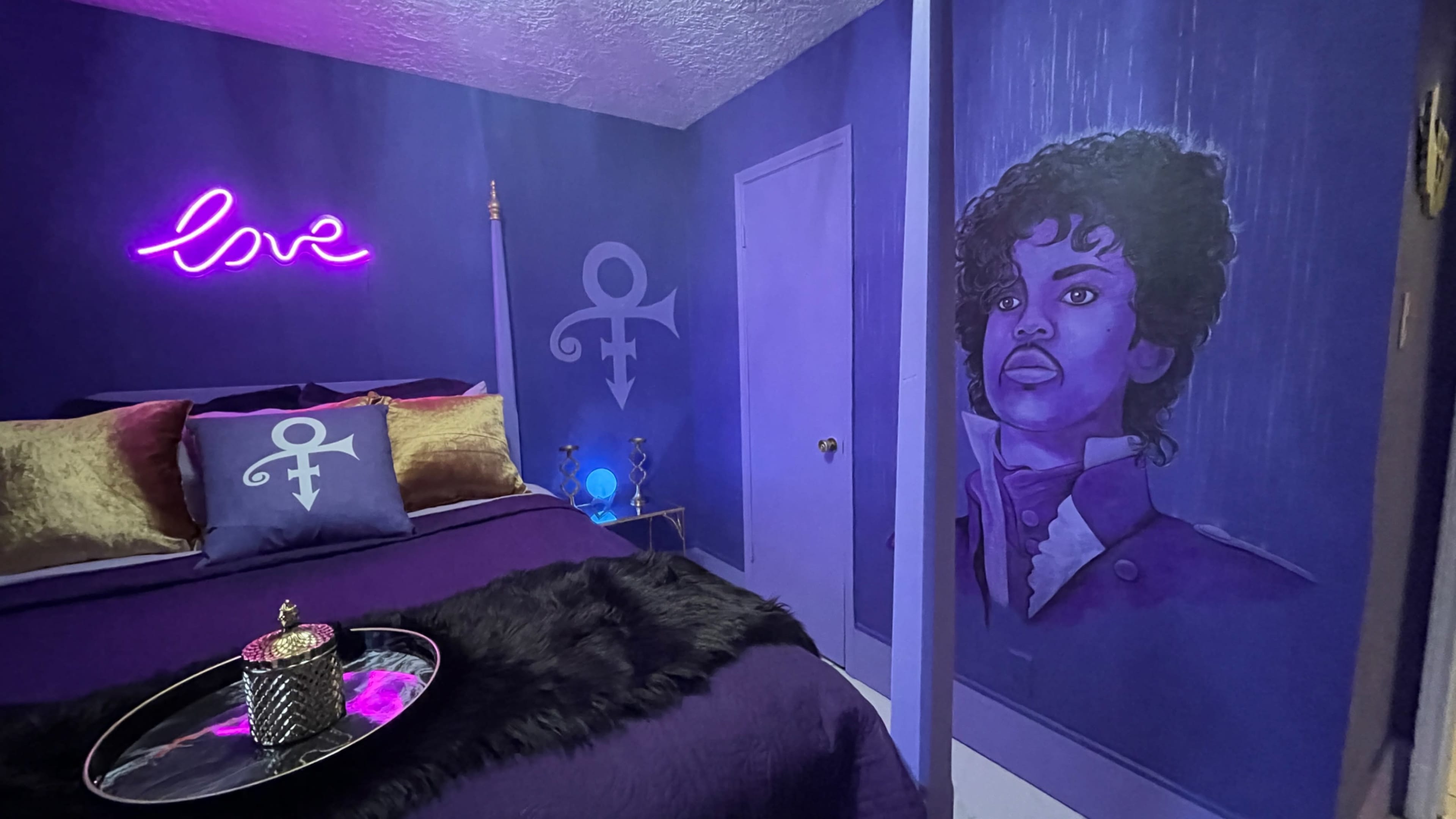 The room features purple walls with a neon "love" sign, a portrait of a person, and decorative pillows with a symbol reminiscent of Prince's emblem.