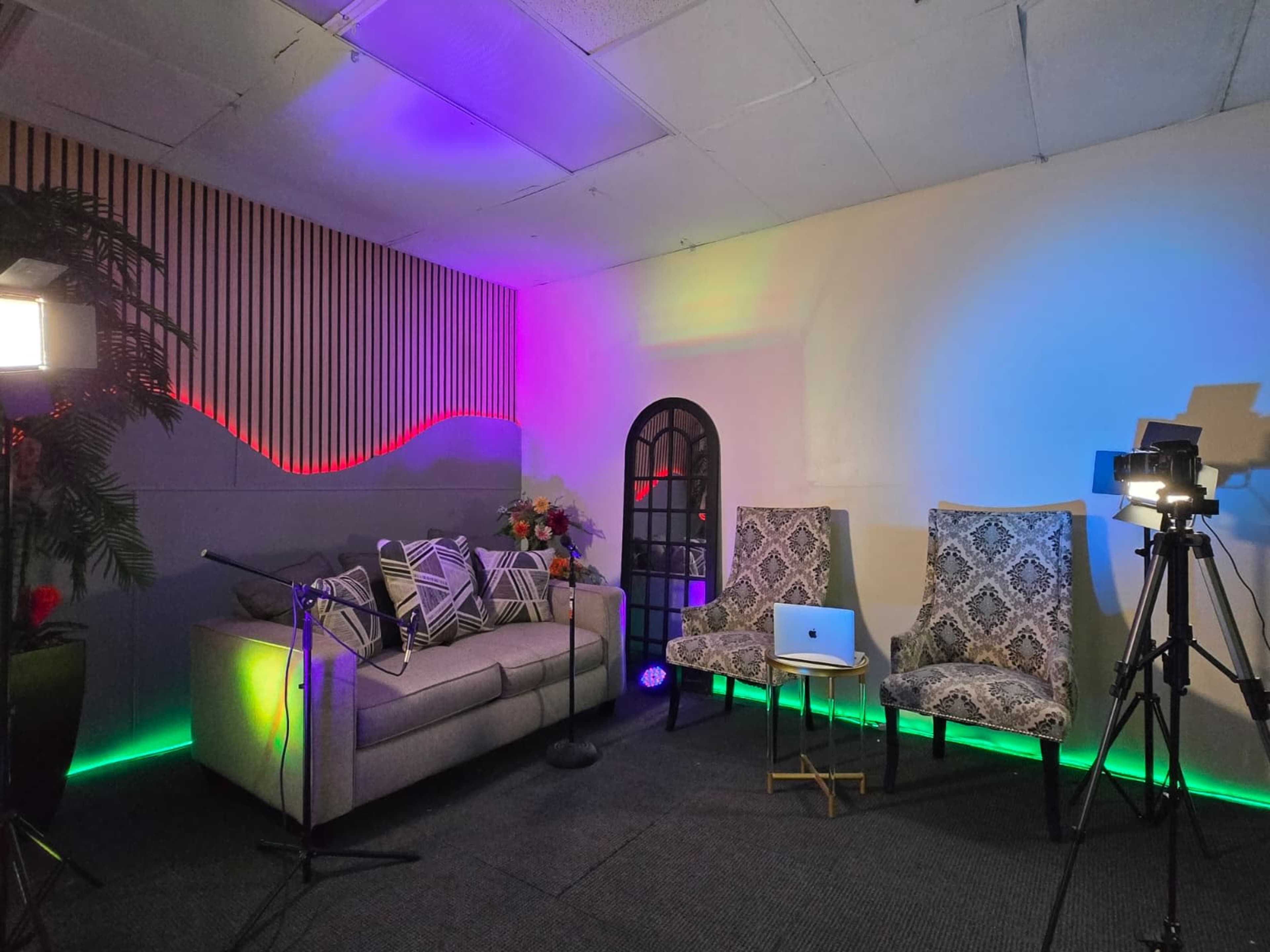 The image shows a brightly lit studio setup featuring a sofa adorned with decorative pillows, two patterned chairs, and professional camera equipment, all set against a colorful backdrop.