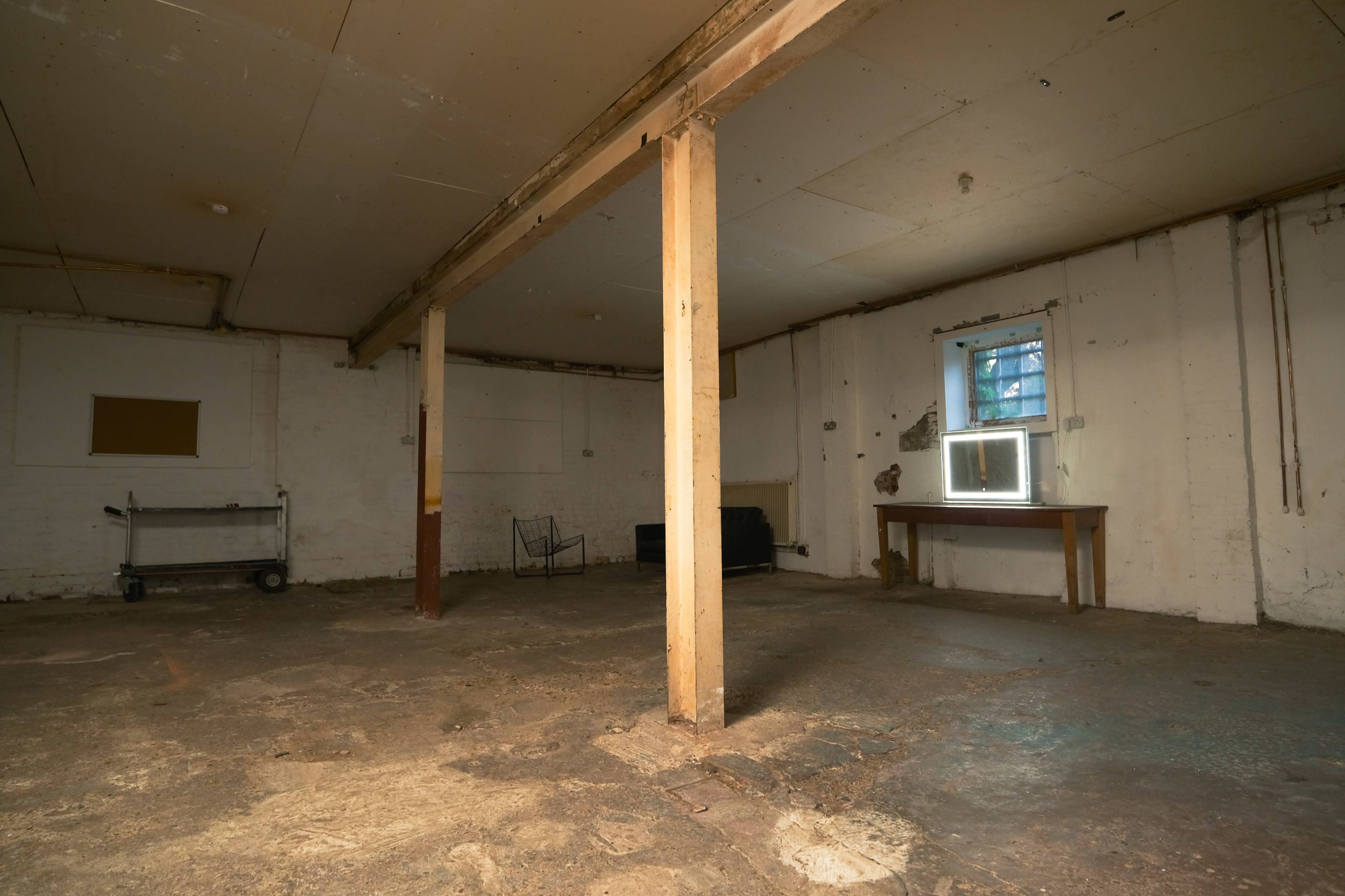 The image shows an empty basement room with a concrete floor, wooden support beams, and minimal furnishings, including a table and a window.