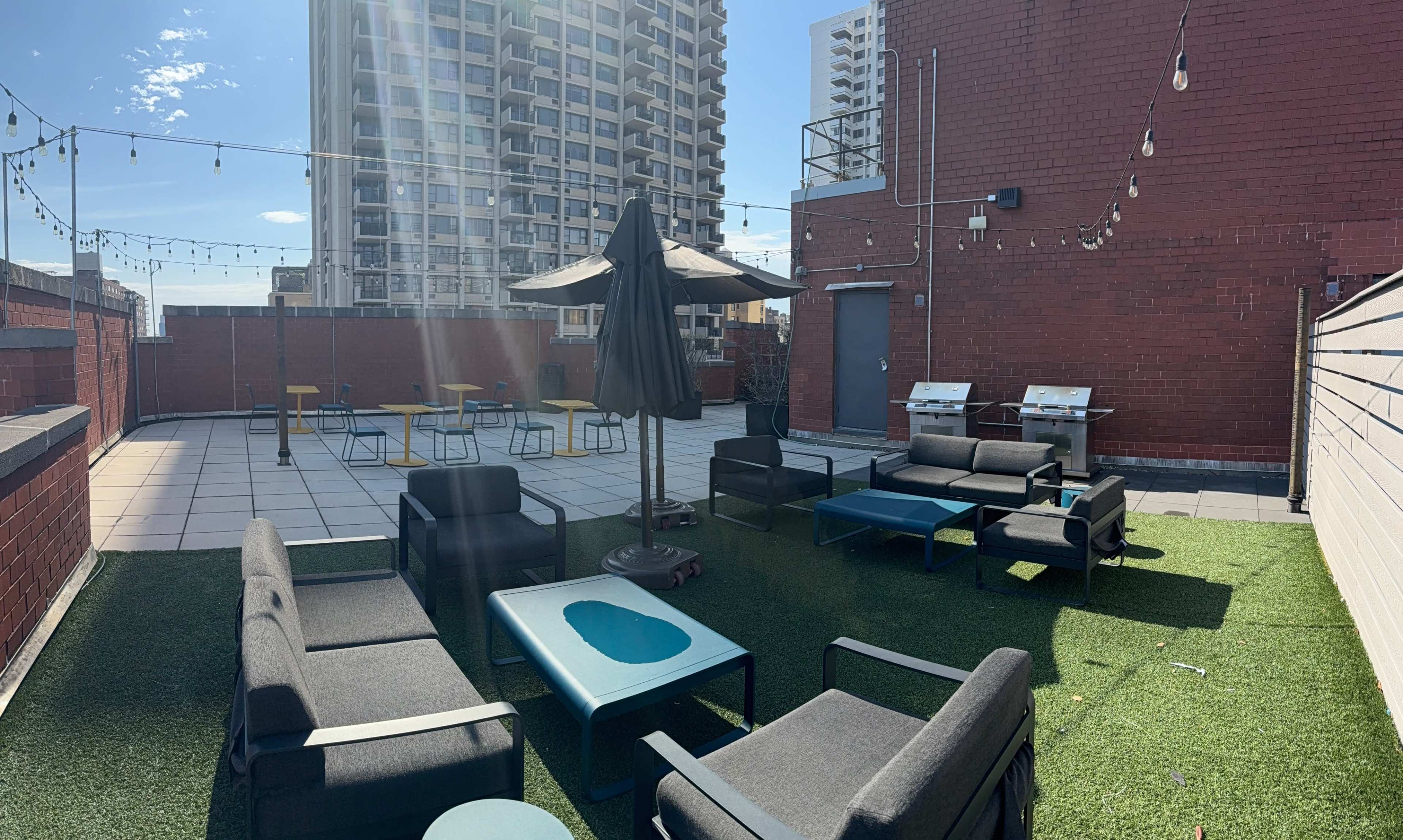 The image shows a rooftop patio with modern seating, green turf, and a grill area, surrounded by tall buildings under a clear blue sky.