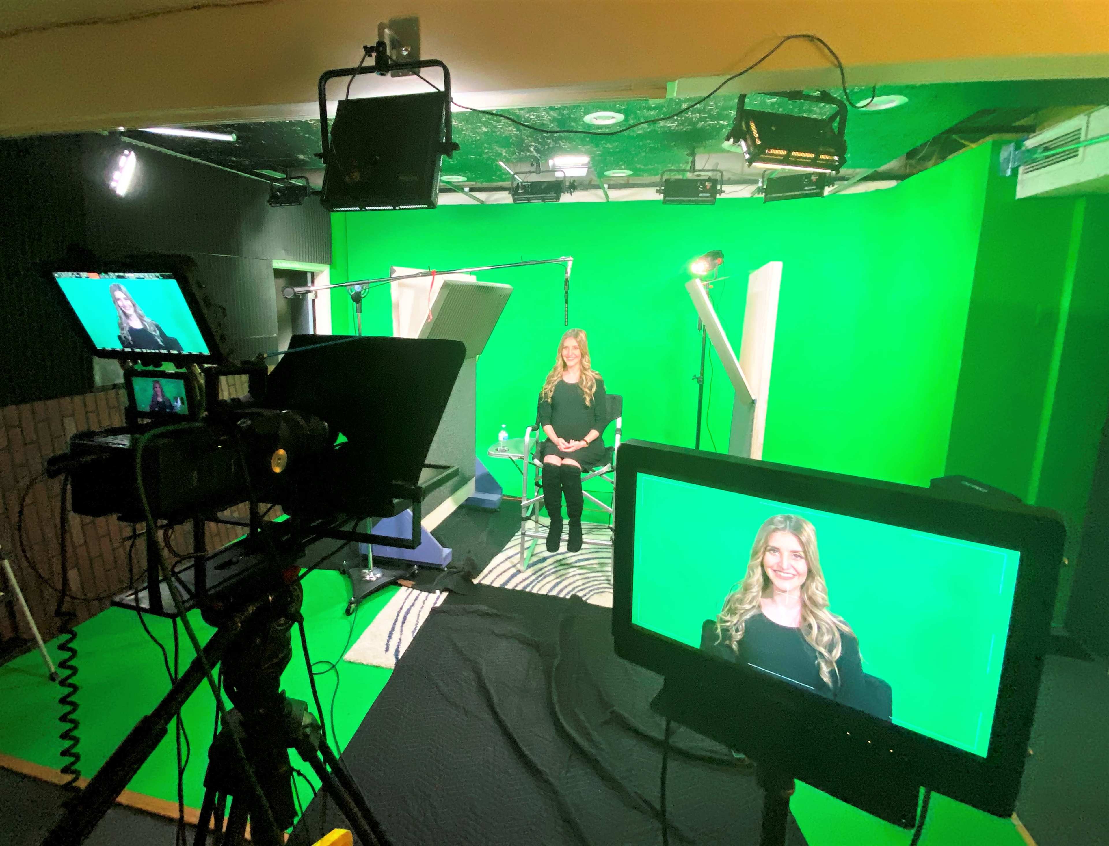 The image shows a woman seated in front of a green screen, surrounded by cameras and lighting equipment in a studio setting.